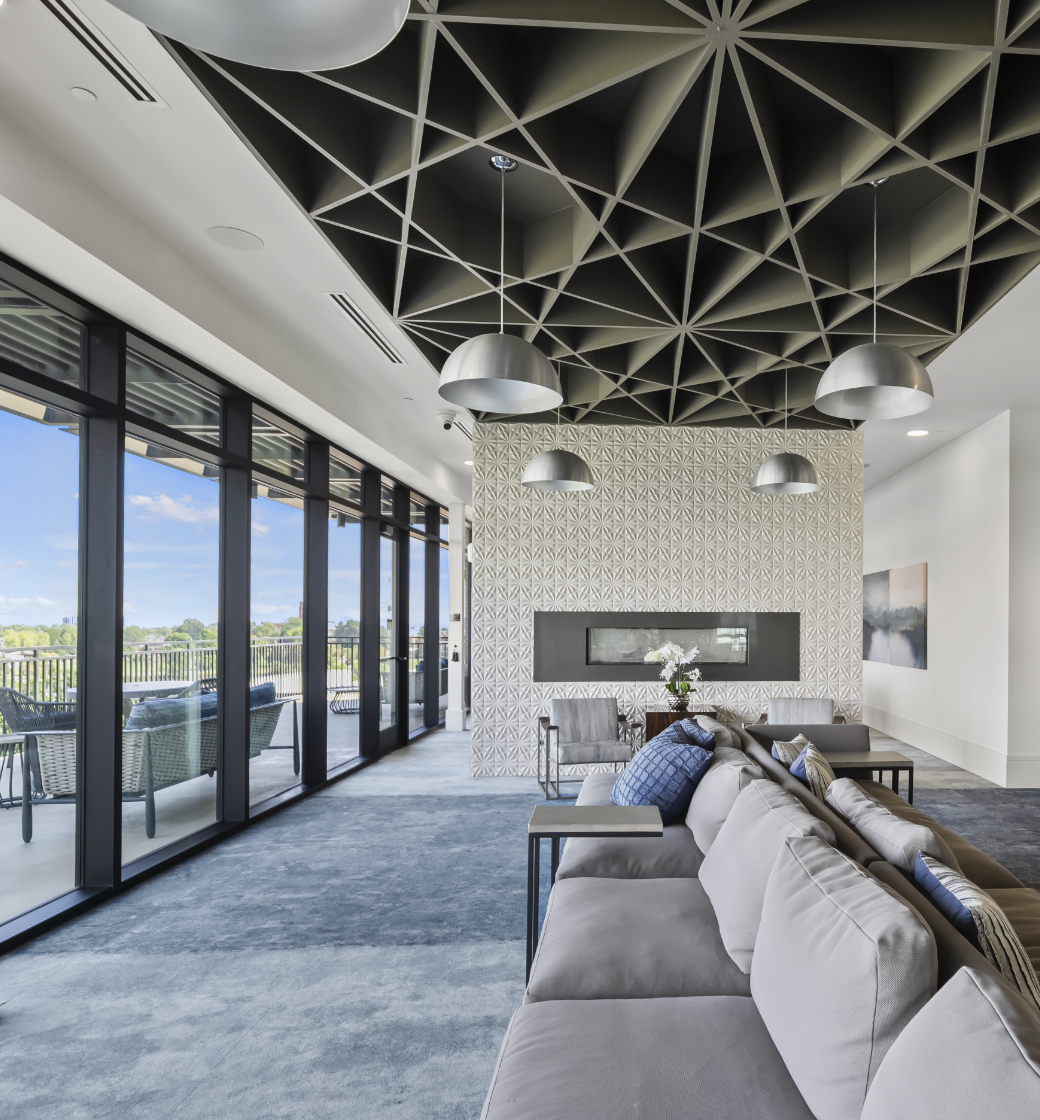 Modern lounge with large windows, blue carpet, gray seating, geometric black ceiling, and a white fireplace mantel.