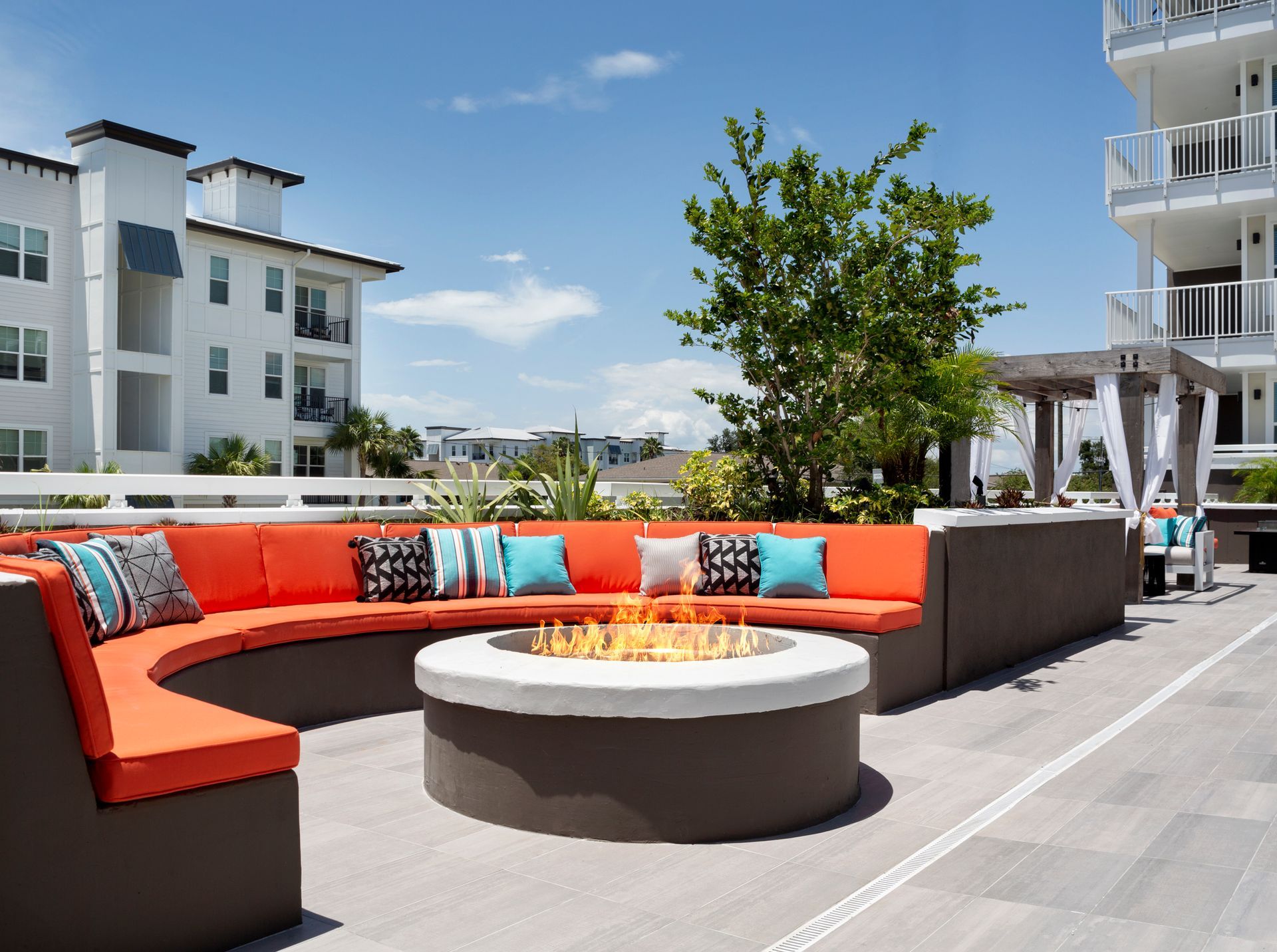Rooftop lounge with orange curved seating around a fire pit, overlooking buildings and blue sky.