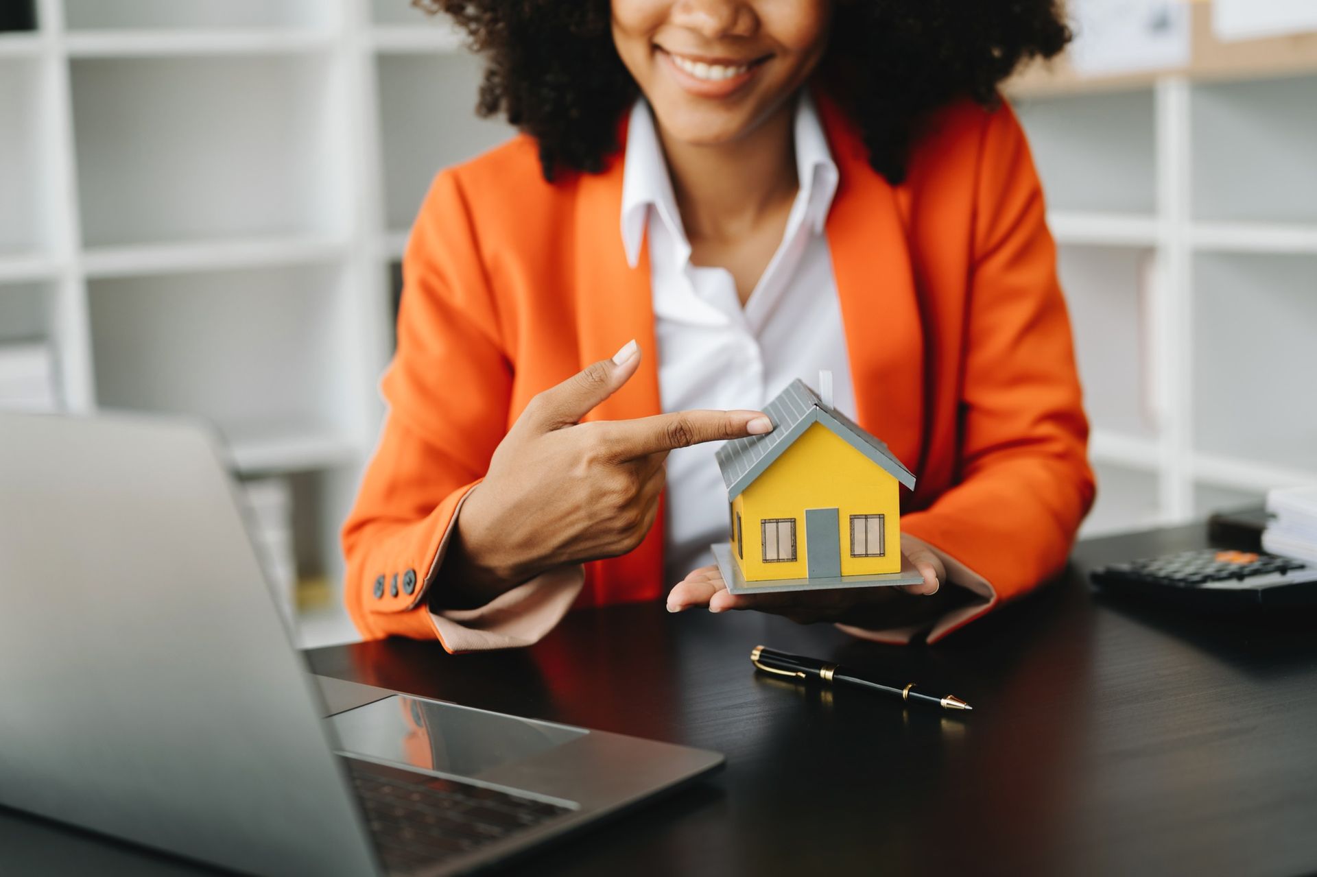 Woman in orange blazer pointing at a small house model on a desk beside a laptop.