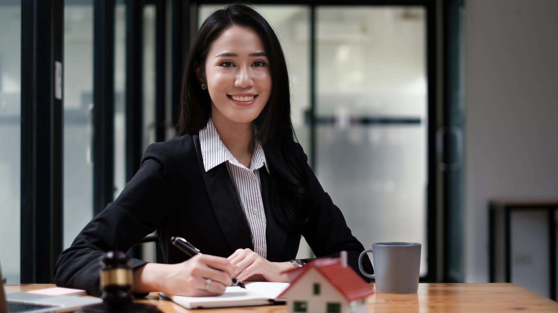Woman in a black blazer smiles at a desk, writing beside a mug and small house model in an office.
