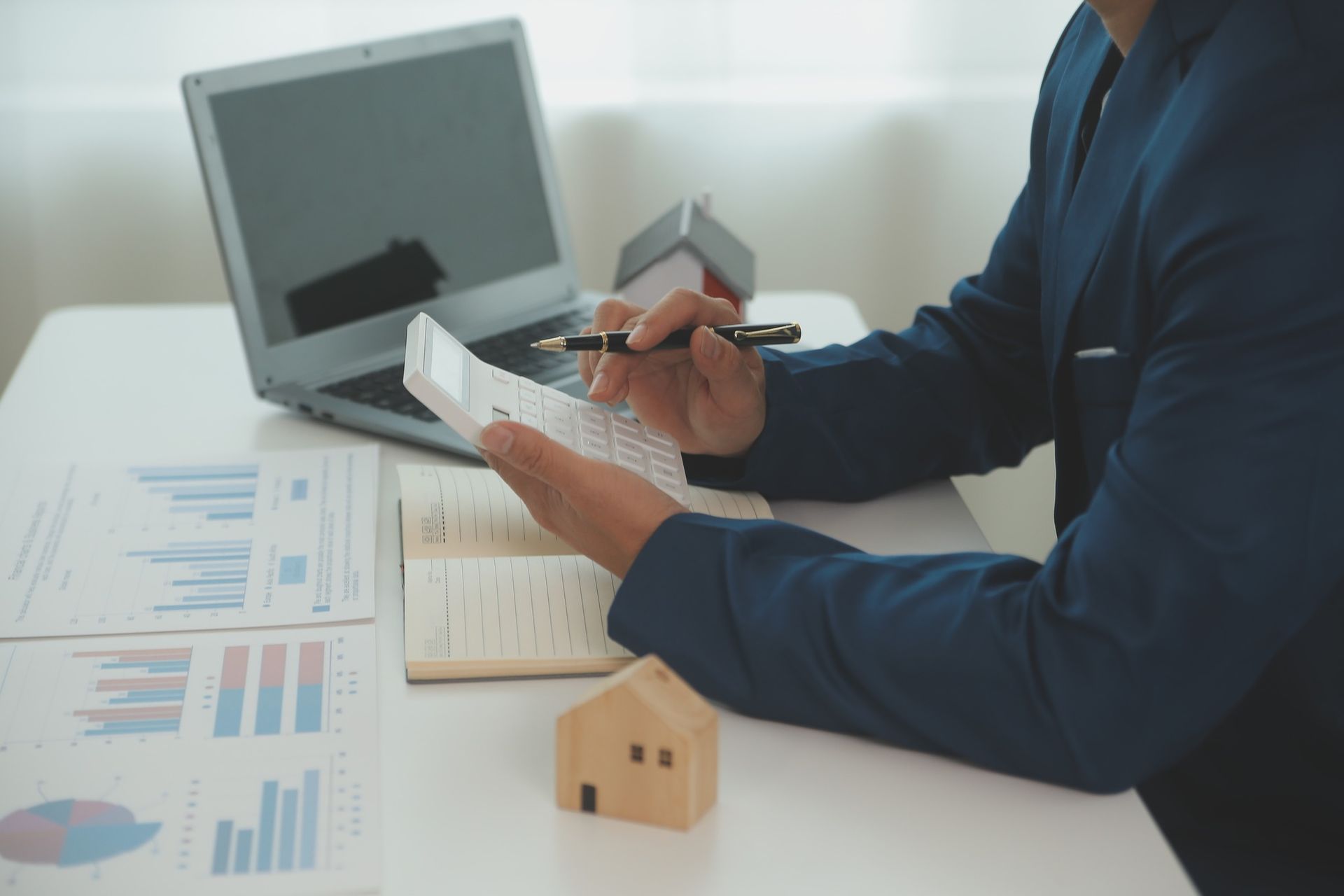 Person reviewing documents at a desk with a laptop, charts, notebook, and small house model