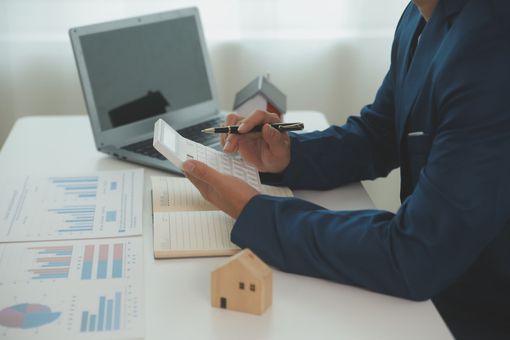 Person reviewing financial documents at a desk with a laptop and calculator, beside a small house model