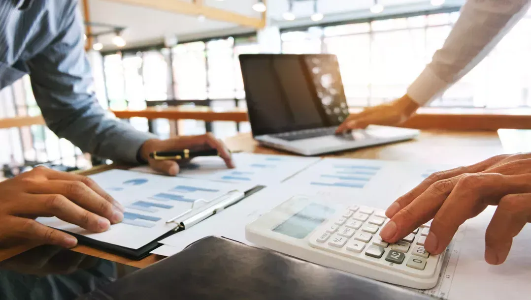 Hands using a calculator and tablet at a bright office desk with a laptop in the background