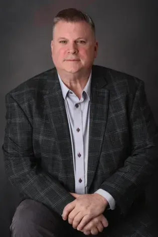 Portrait of a seated man in a dark plaid blazer and light blue shirt against a gray background
