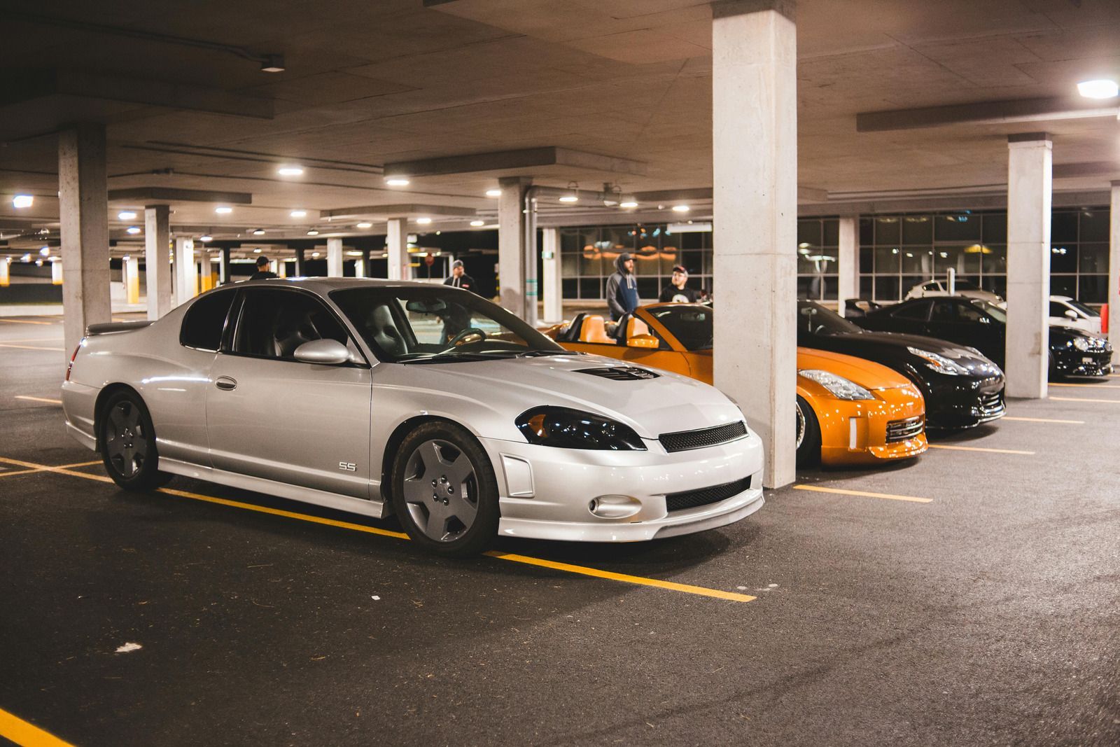 Three sports cars parked in an underground garage, including a silver car in front and orange cars behind.