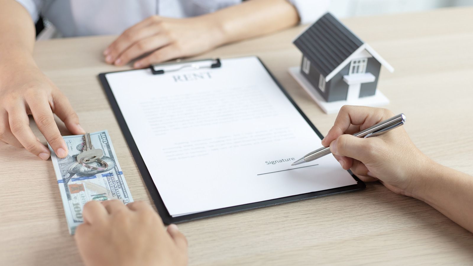 Hands signing a document beside a small model house and cash on a desk