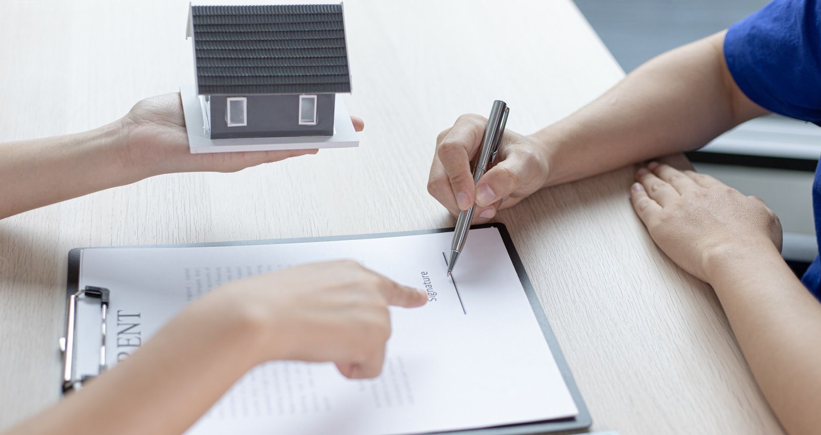 Hands holding a tiny house model over paperwork while another person points on a desk