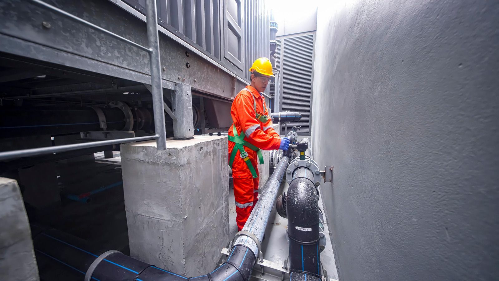 Worker in orange safety gear and yellow hard hat inspecting pipes in a narrow industrial corridor.