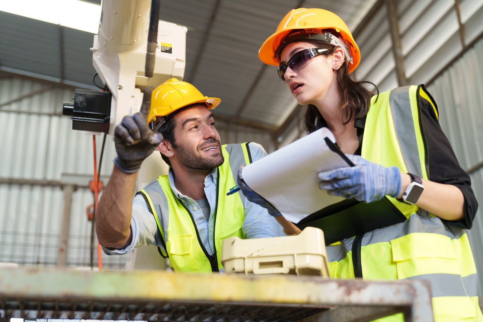 Two construction workers in hard hats and safety vests reviewing plans at a worksite.