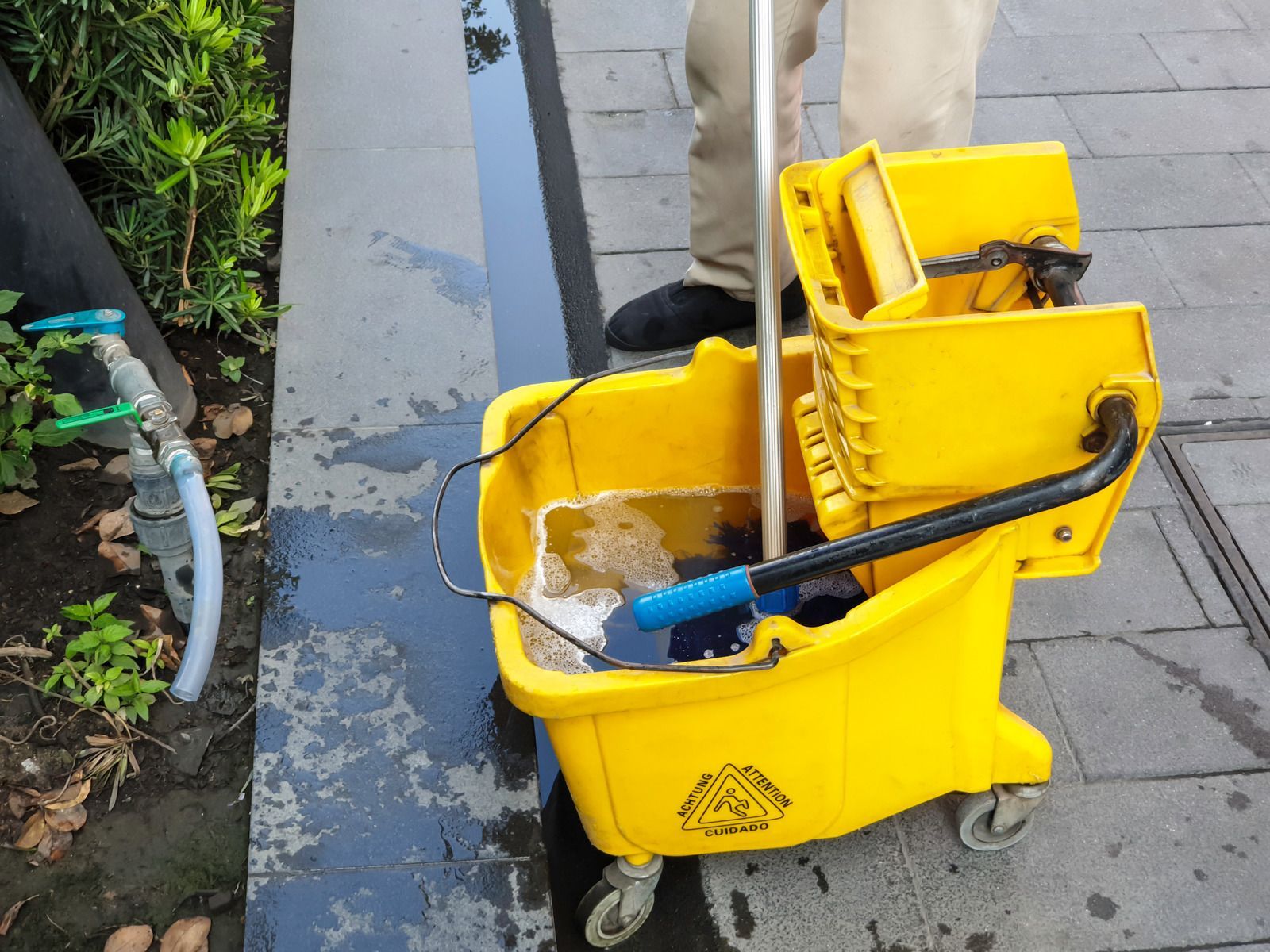 Yellow mop bucket on wheels beside a wet concrete walkway, with a hose and cleaning tools nearby
