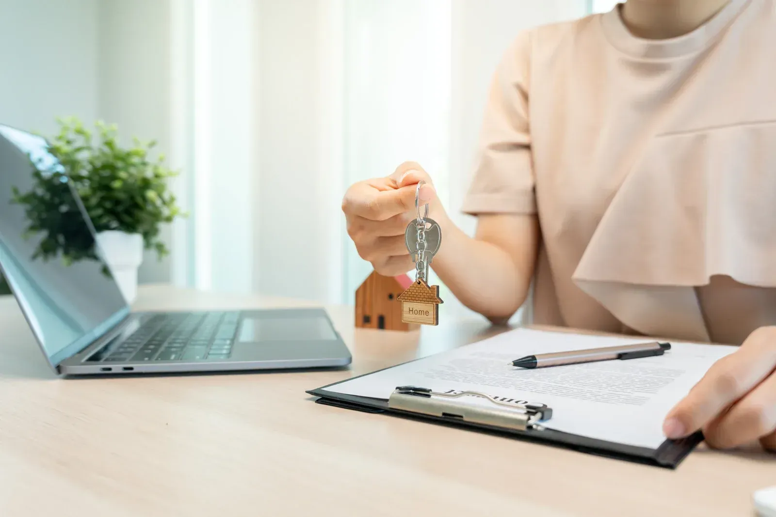 Person holding keys beside a clipboard, laptop, and small house model on a desk