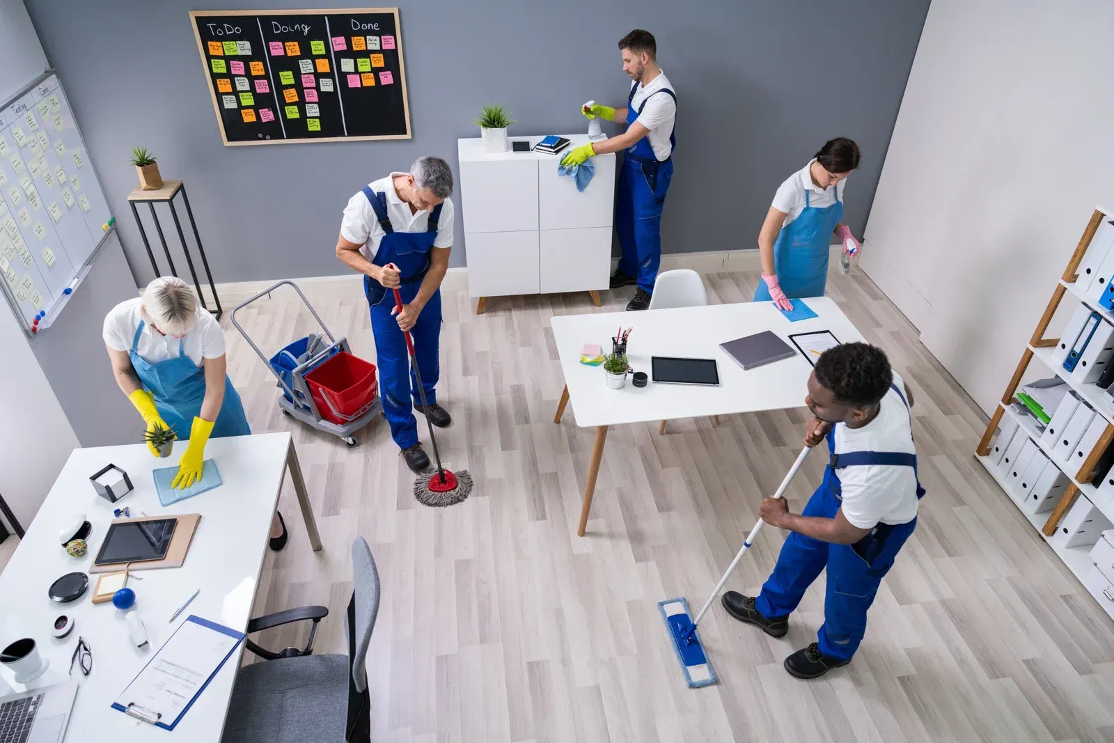 People cleaning a bright office room with mops and supplies around desks and cabinets