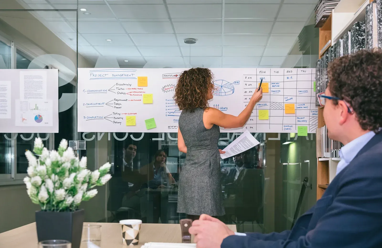 Woman presents notes on glass wall in office meeting, with a seated colleague nearby.