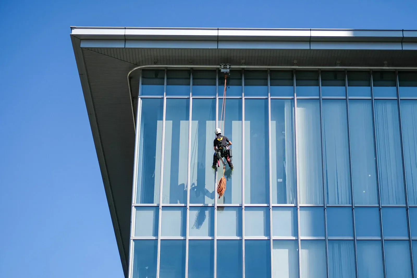 Person suspended by ropes on a glass office building façade under a blue sky