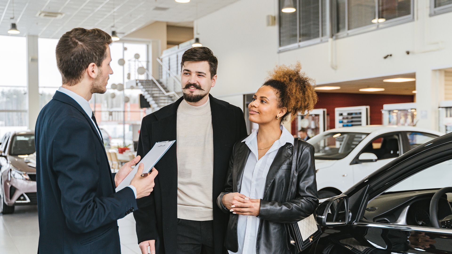 salesman speaking with two customers beside a black car in a bright showroom