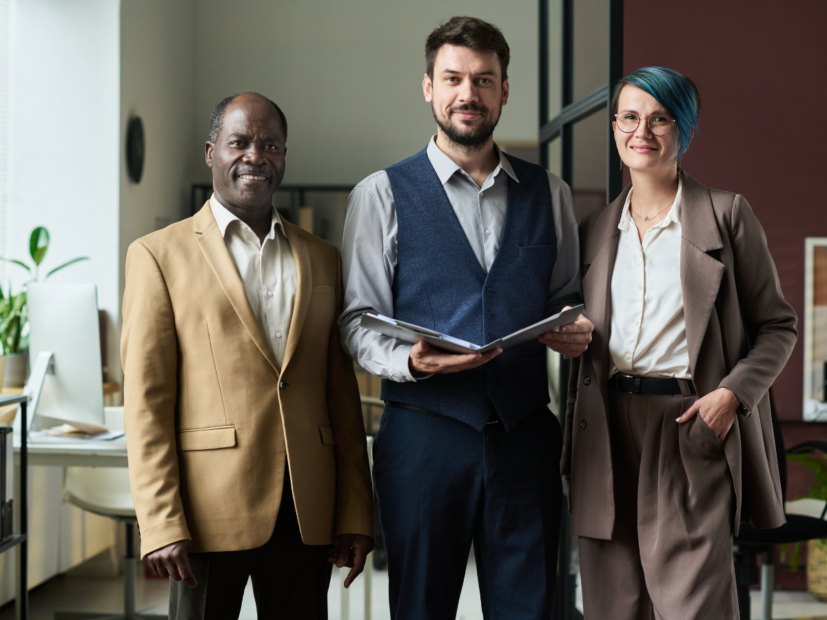 Three coworkers in business attire, smiling at the camera in an office setting.