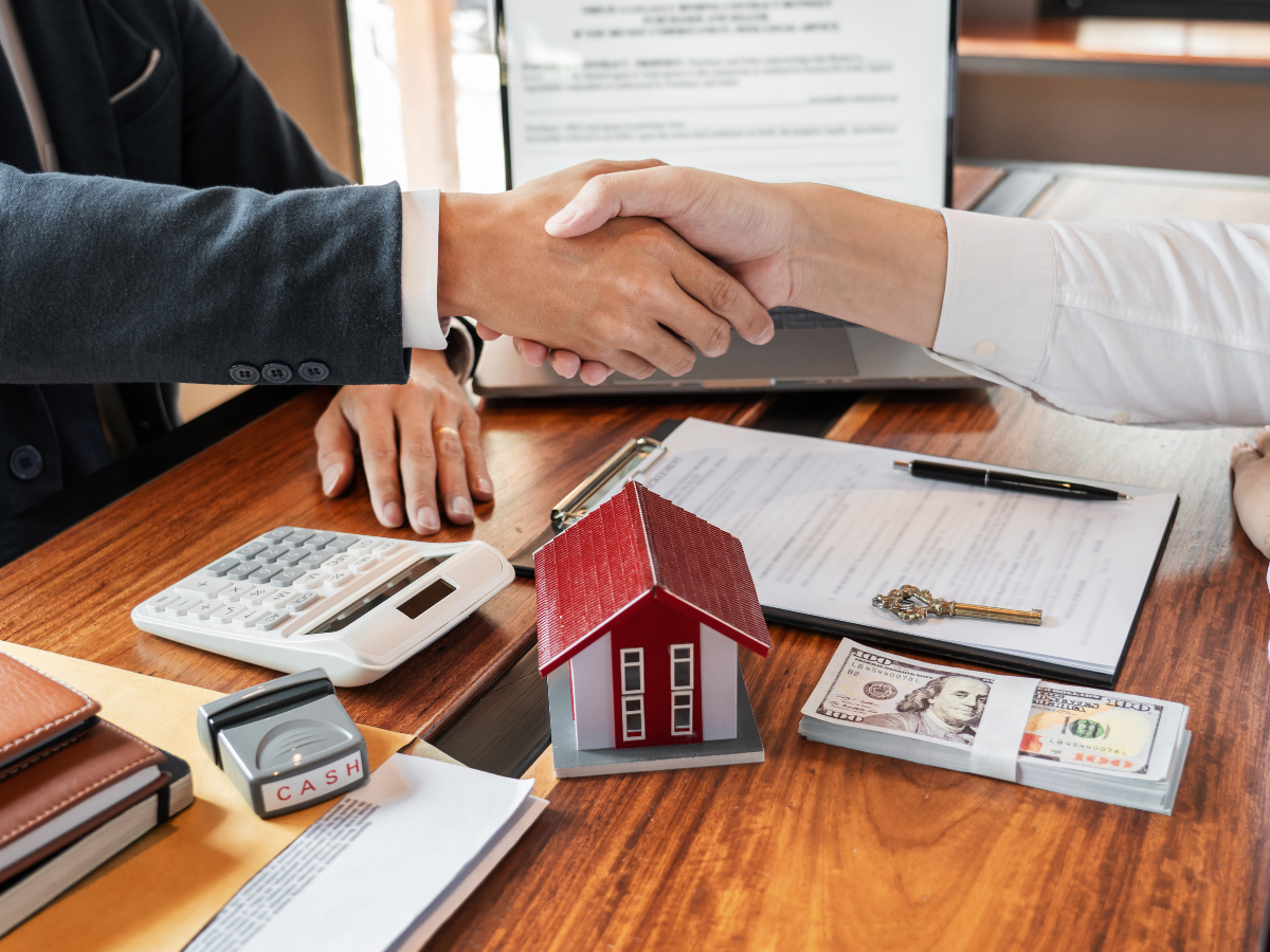 Two people shaking hands over a wooden table with house model, money, and documents.