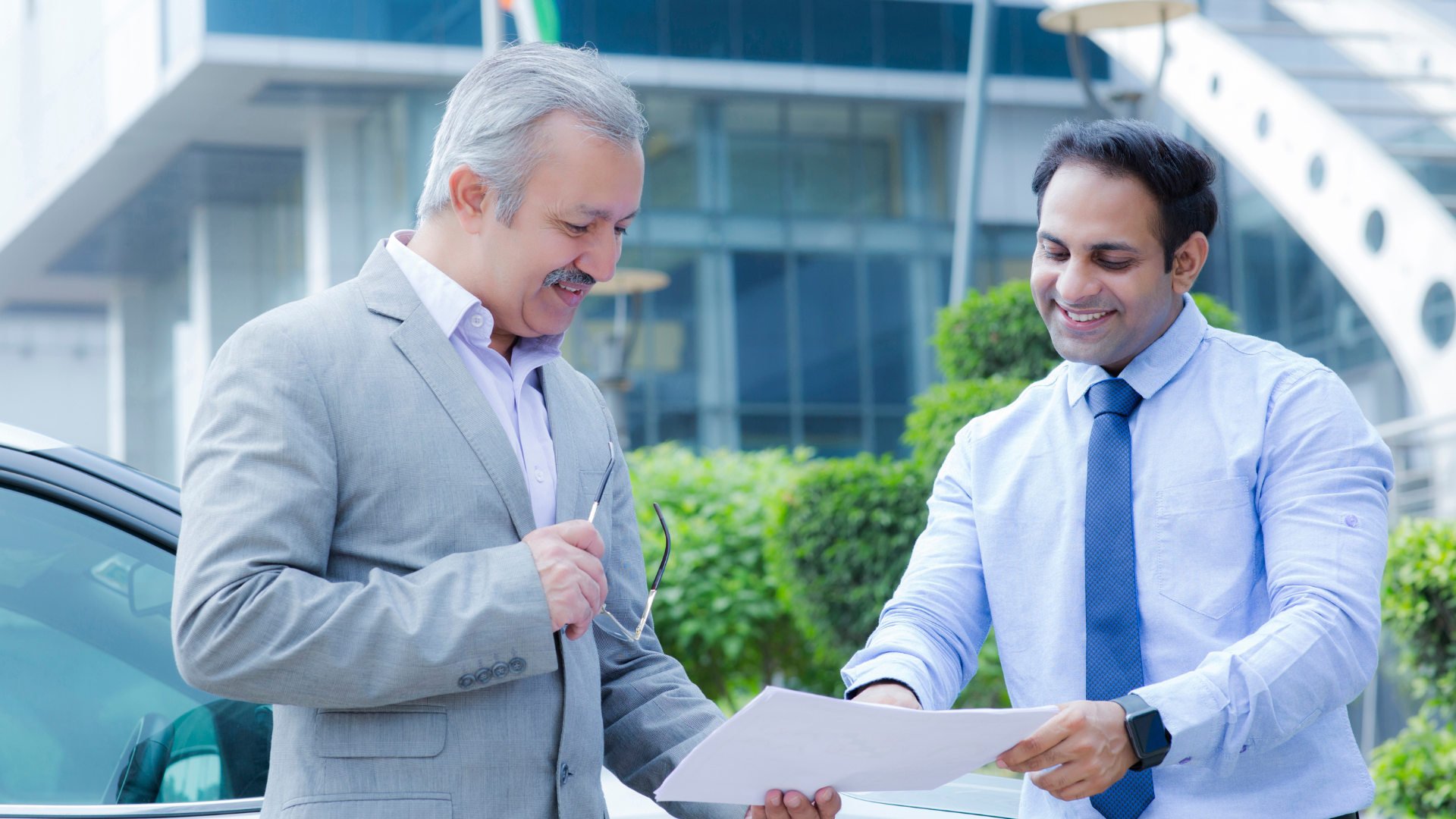 Two businessmen reviewing documents outdoors near a modern office building.