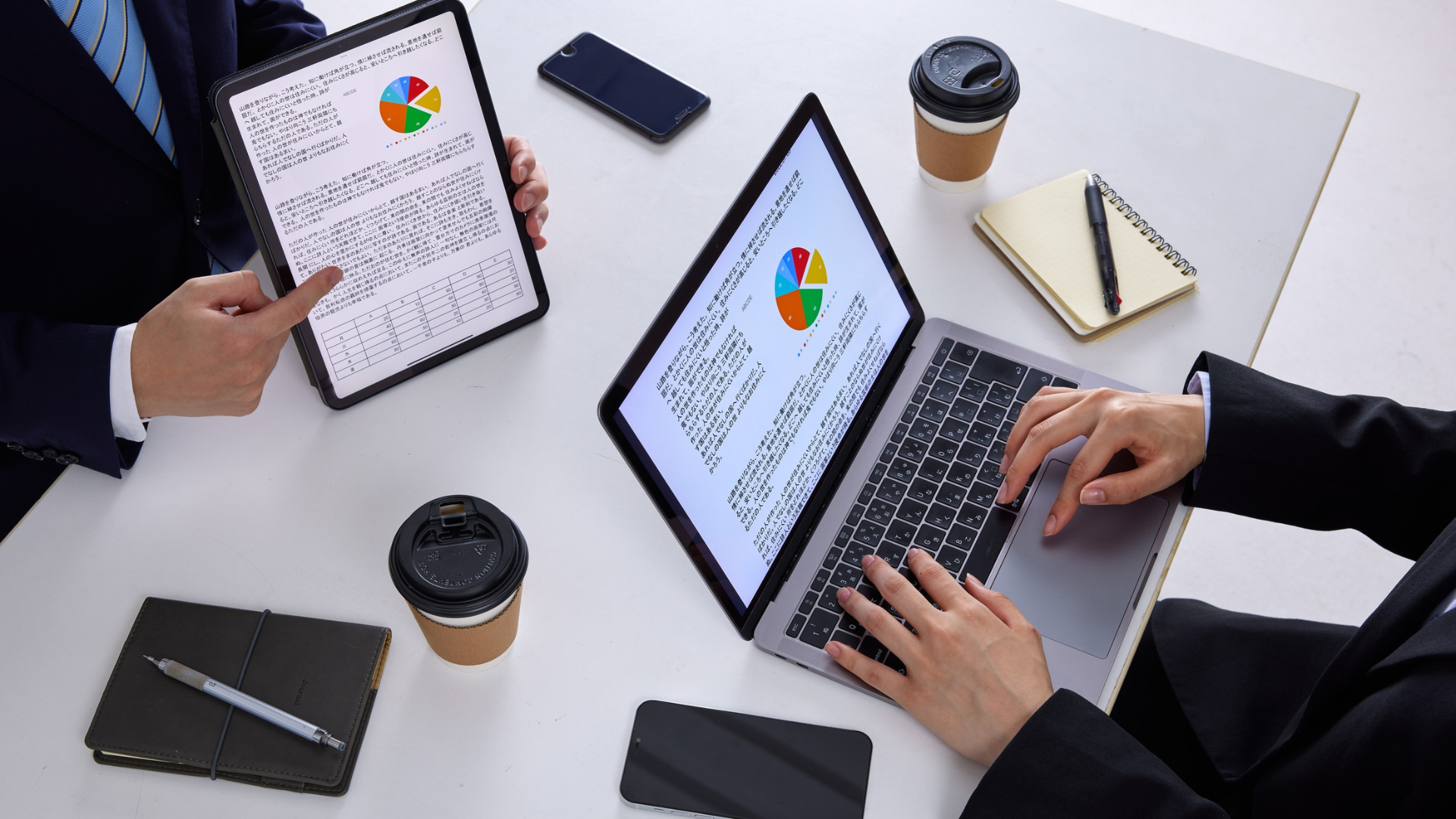 Two people working on laptops at a white desk with coffee cups and notebooks, viewing charts on screens