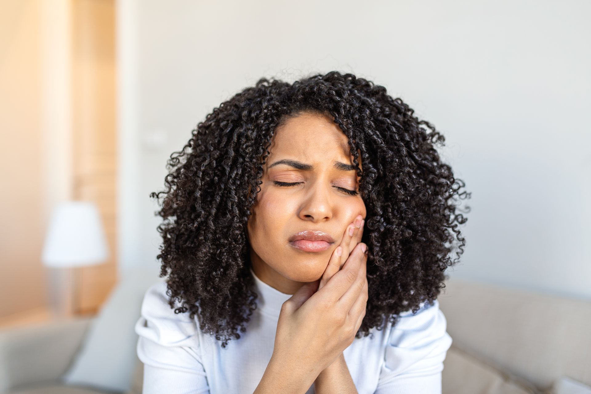 Person sitting indoors, eyes closed, holding a cheek in discomfort on a couch.