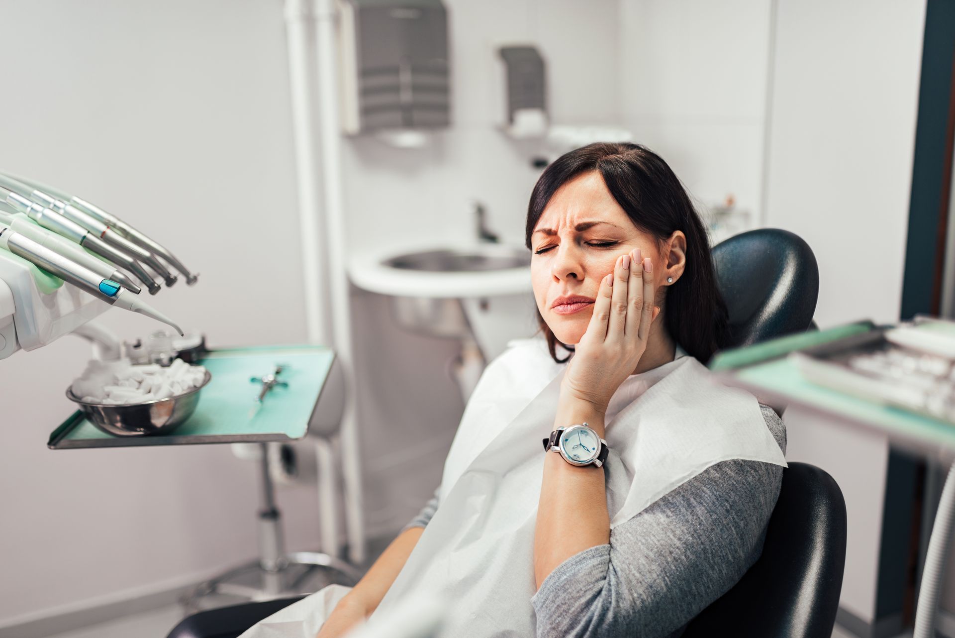 Woman holding her cheek in a dental chair, looking uncomfortable during a dental exam, with tools nearby.