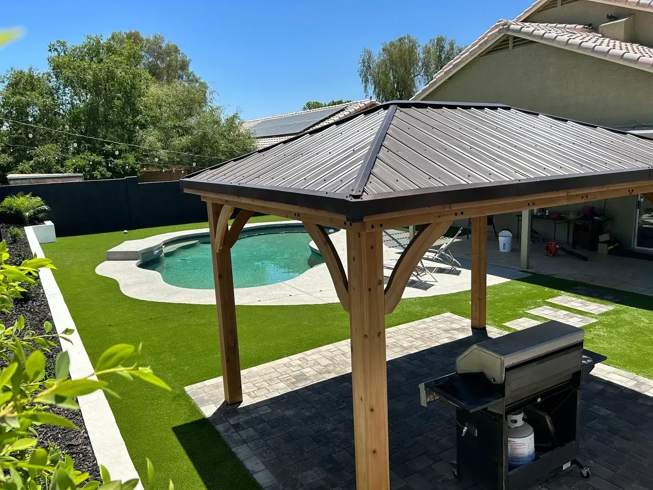 A gazebo with a grill underneath it in a backyard with a pool.