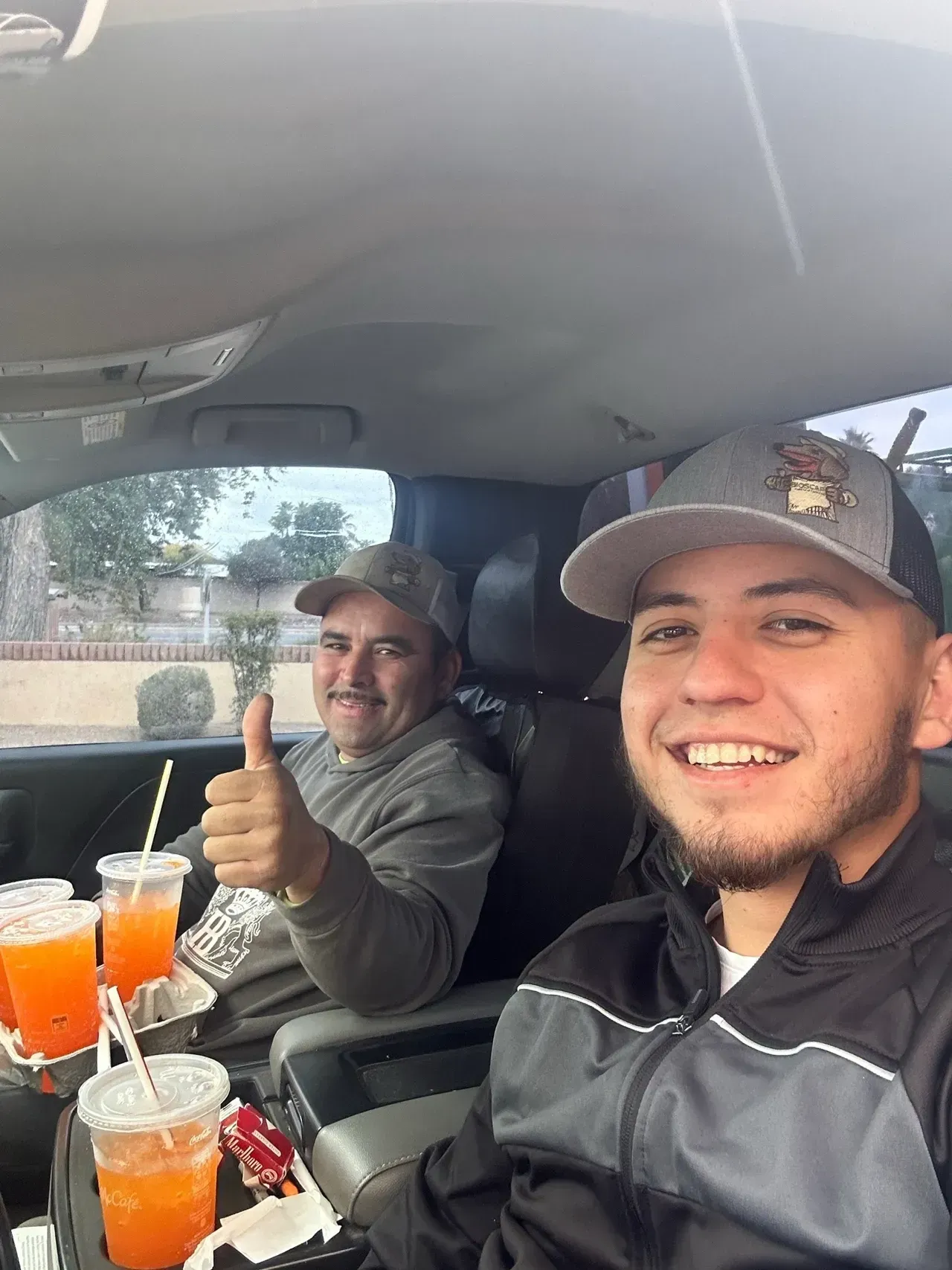 Two men are sitting in a car drinking orange juice and giving a thumbs up.