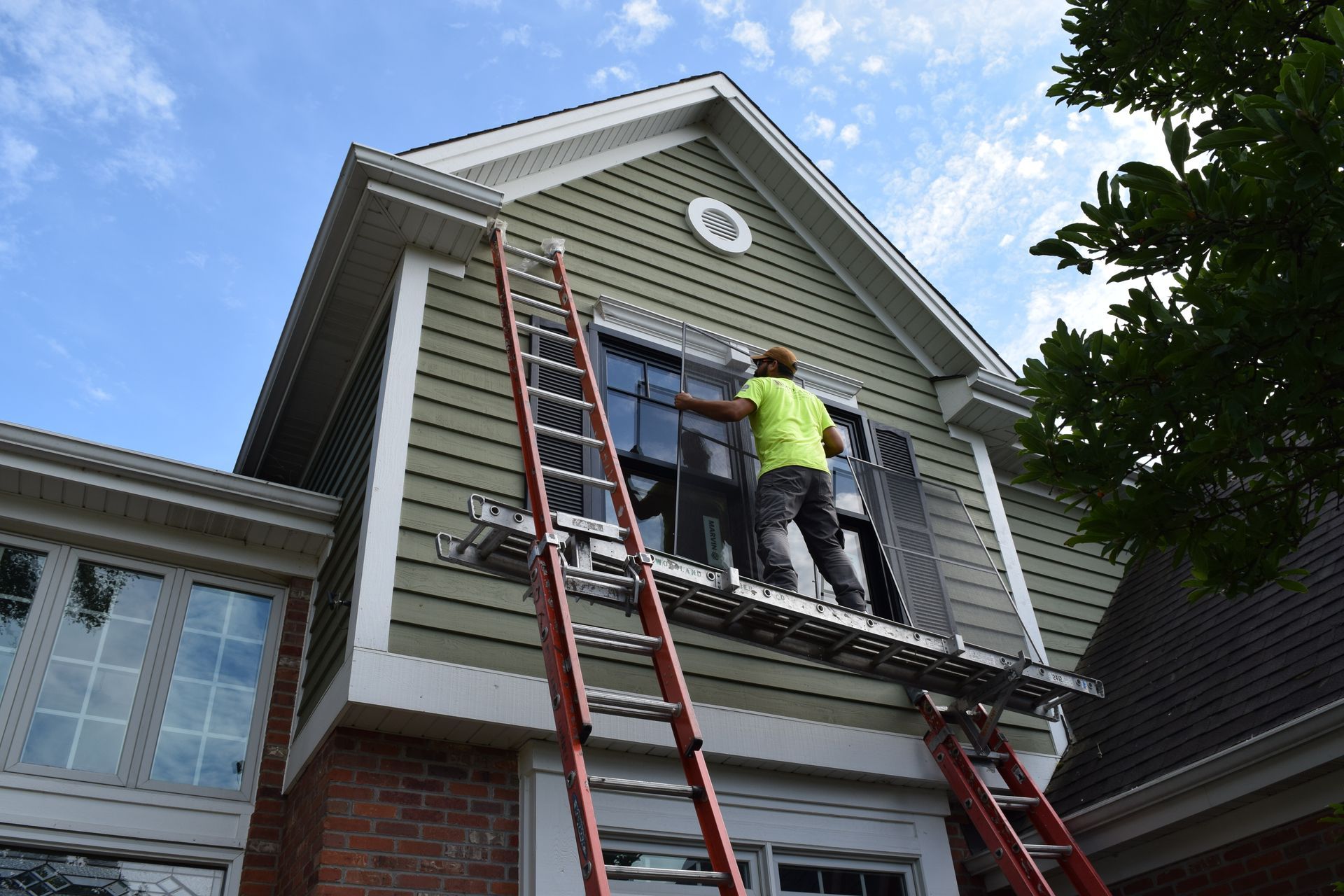 Person on a ladder washing windows of a two-story house with green siding and a brick base.