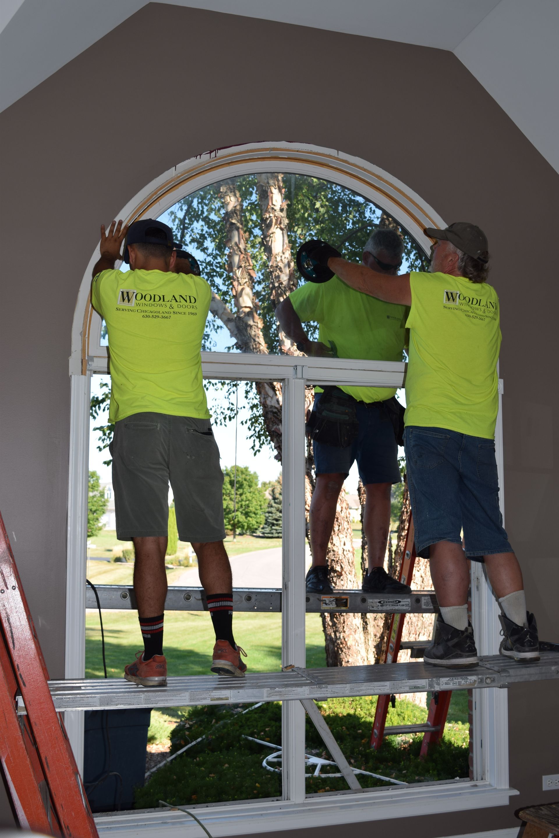 Three people wearing yellow shirts installing a white, arched window in a room with tan walls.