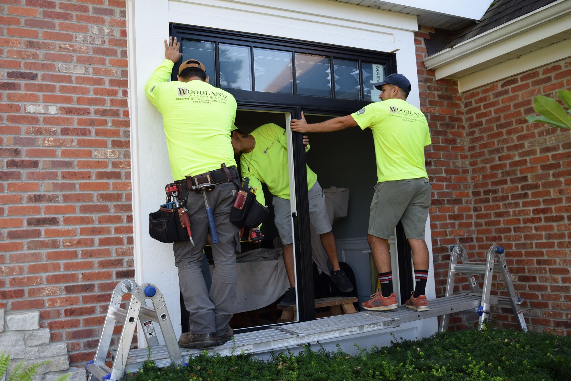 Three workers in neon shirts installing a black framed window into a brick building.