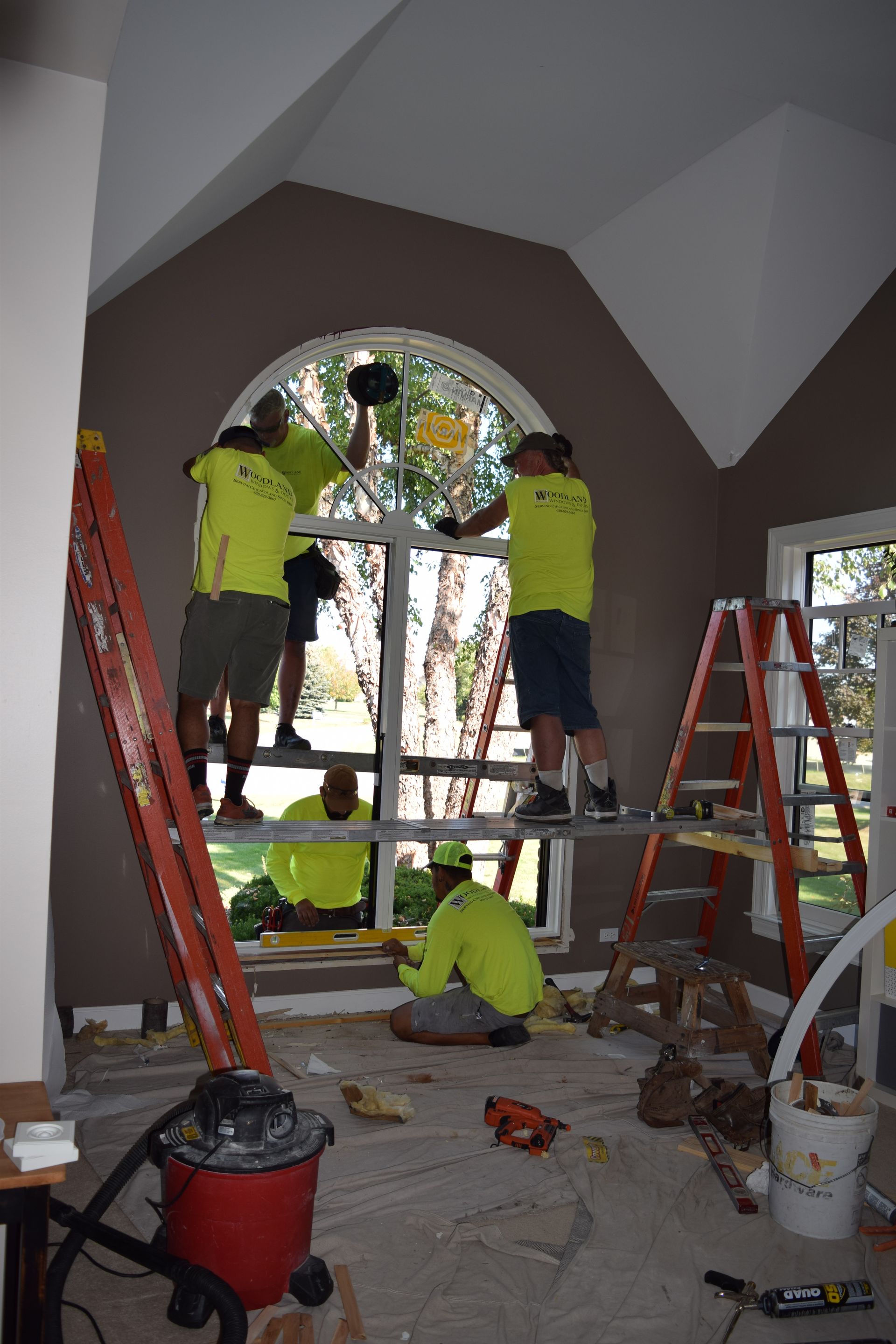 Workers in yellow shirts replacing a large arched window inside a room.
