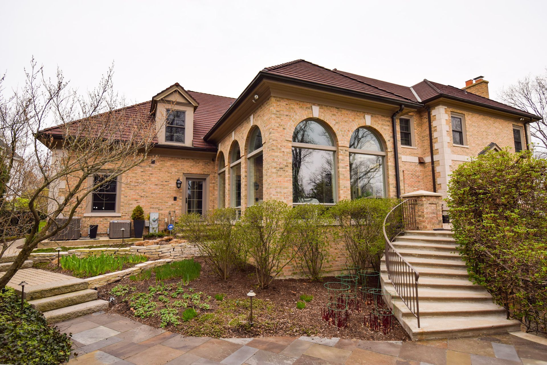 Two-story brick house with arched windows, red tile roof, and stone stairs leading up from the yard.