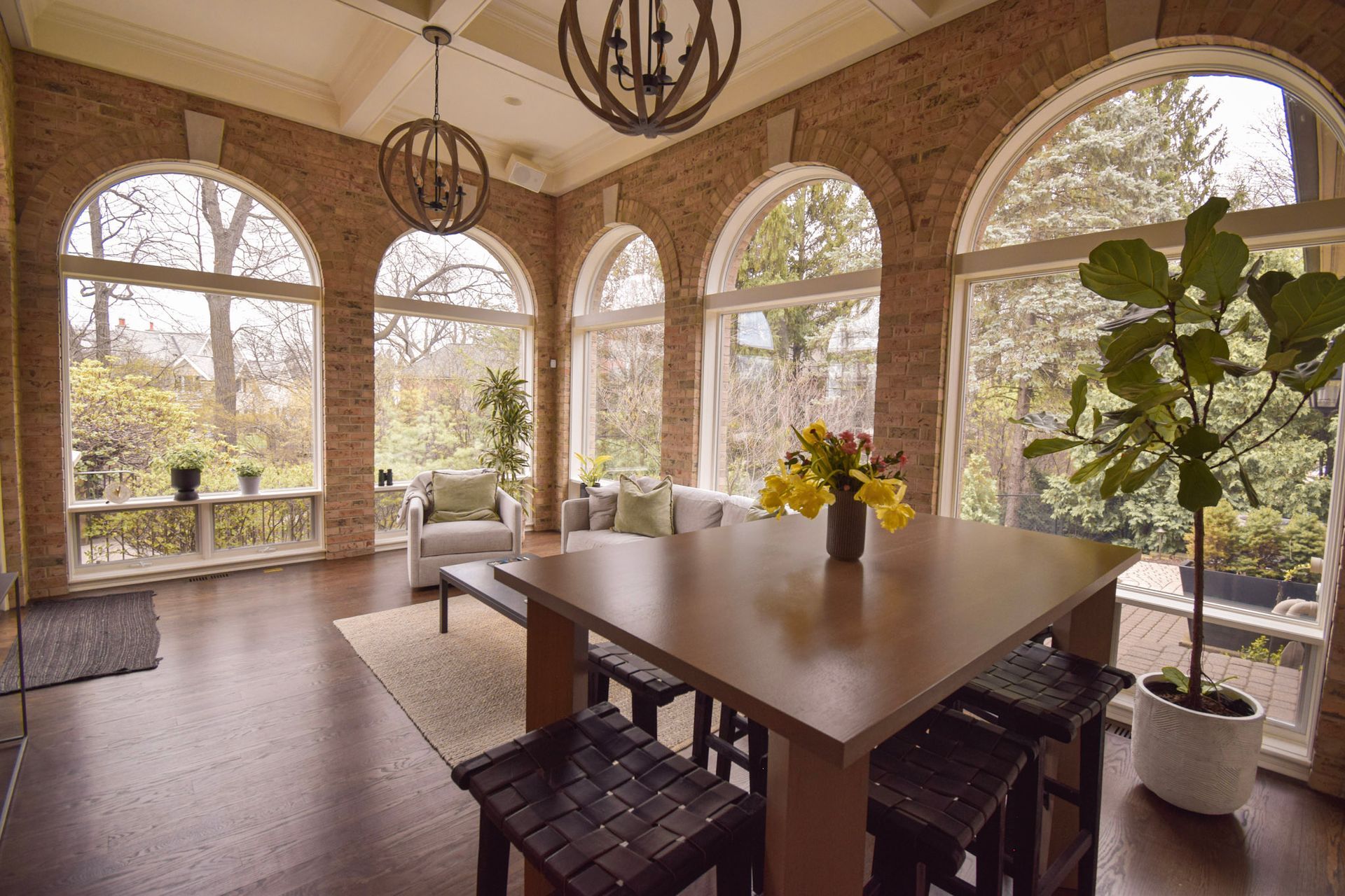 Sunroom with arched windows, dining table, and sofa, surrounded by greenery, warm brick walls.