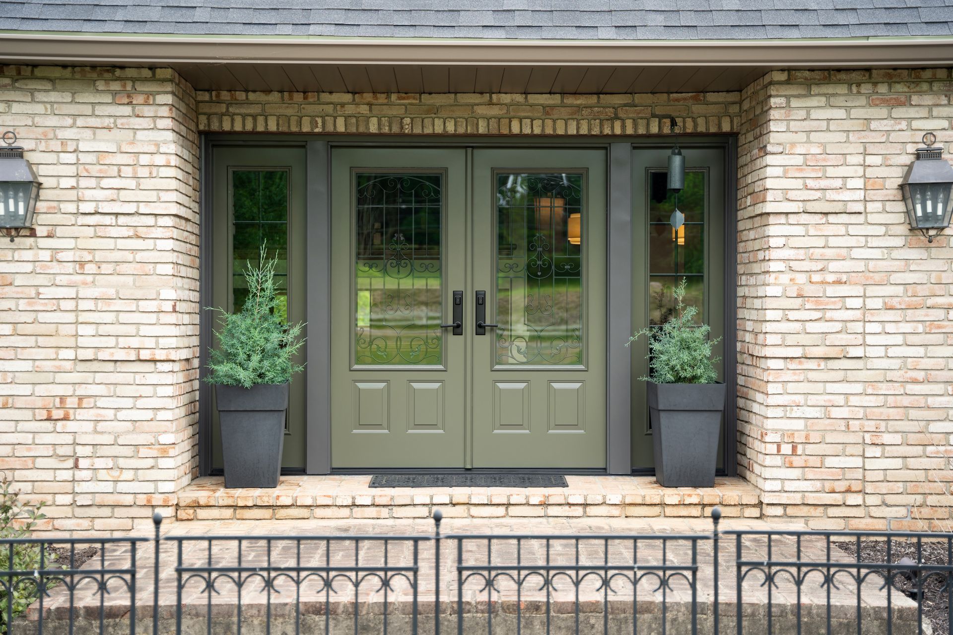 The front door of a brick house with green doors and potted plants.