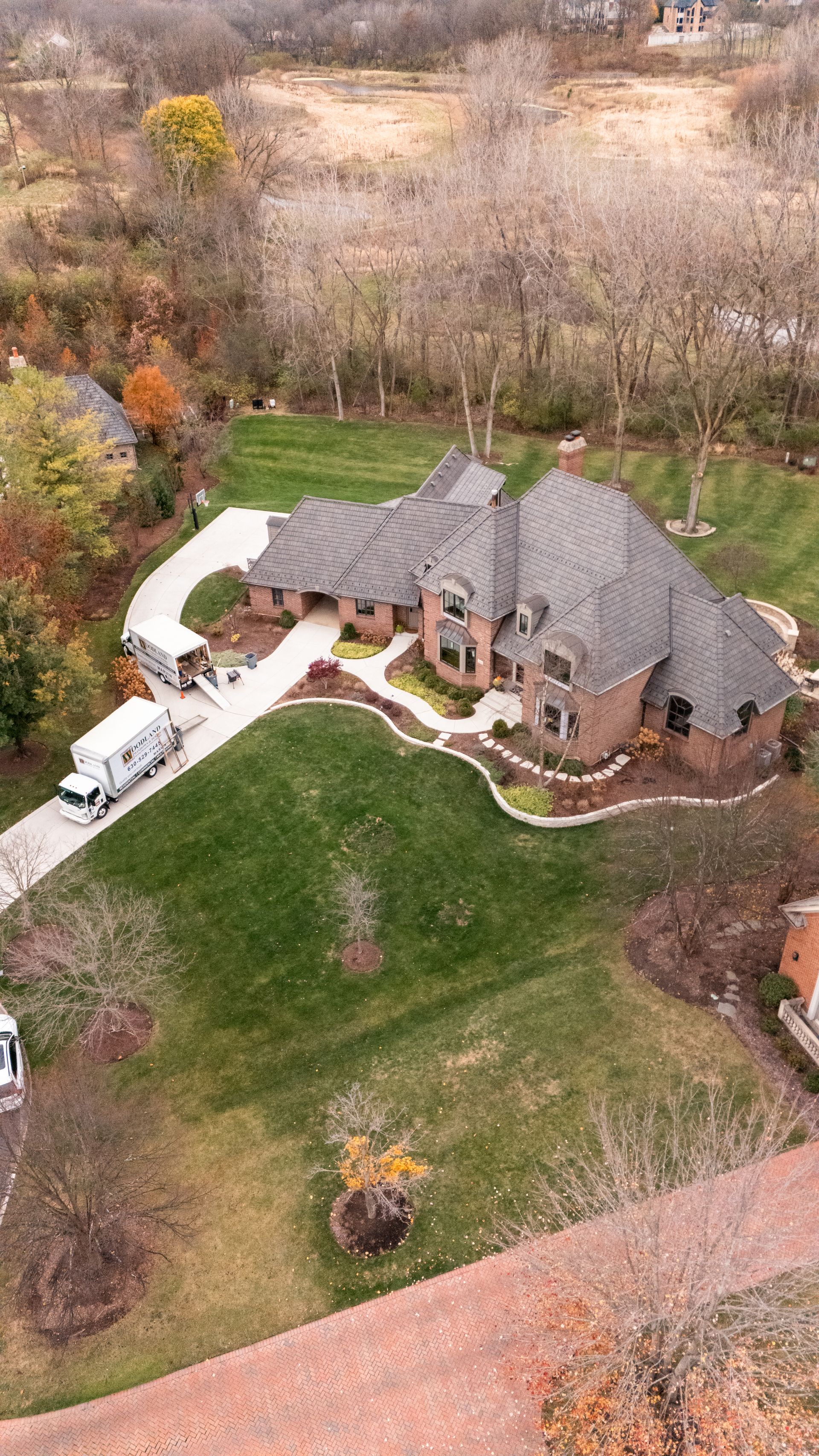 Large brick house with curved driveway, moving truck parked outside. Green lawn, fall foliage.