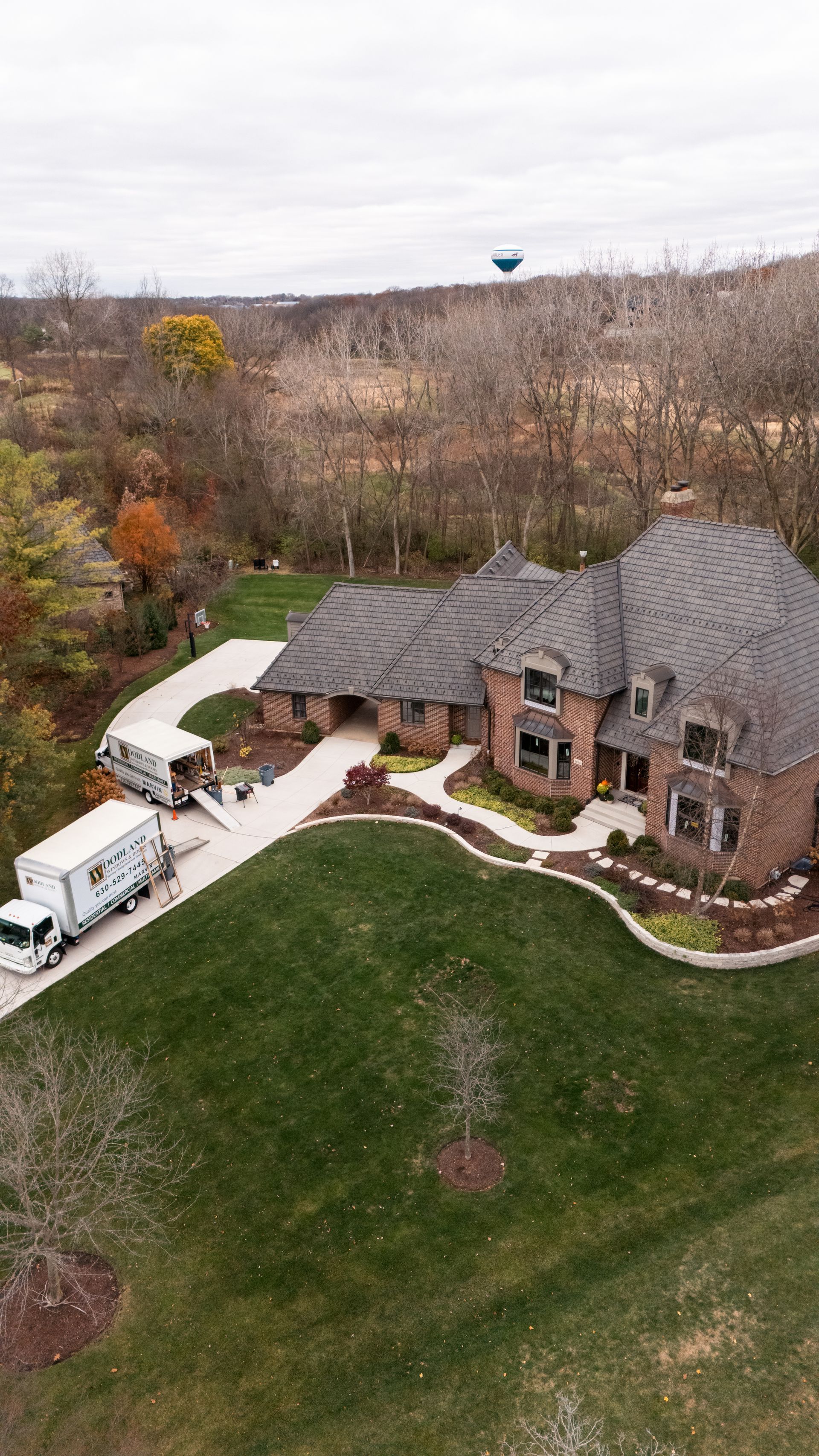 Two moving trucks parked in driveway of large brick house with green lawn; autumn foliage in background.