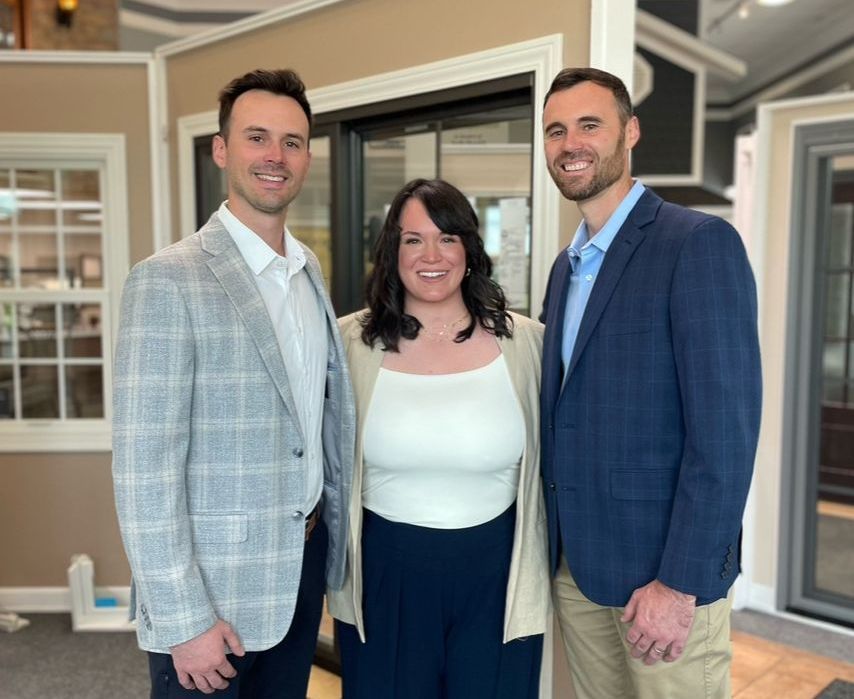 Three people, two men and a woman, smiling in a showroom. Men wear blazers, woman a cardigan.