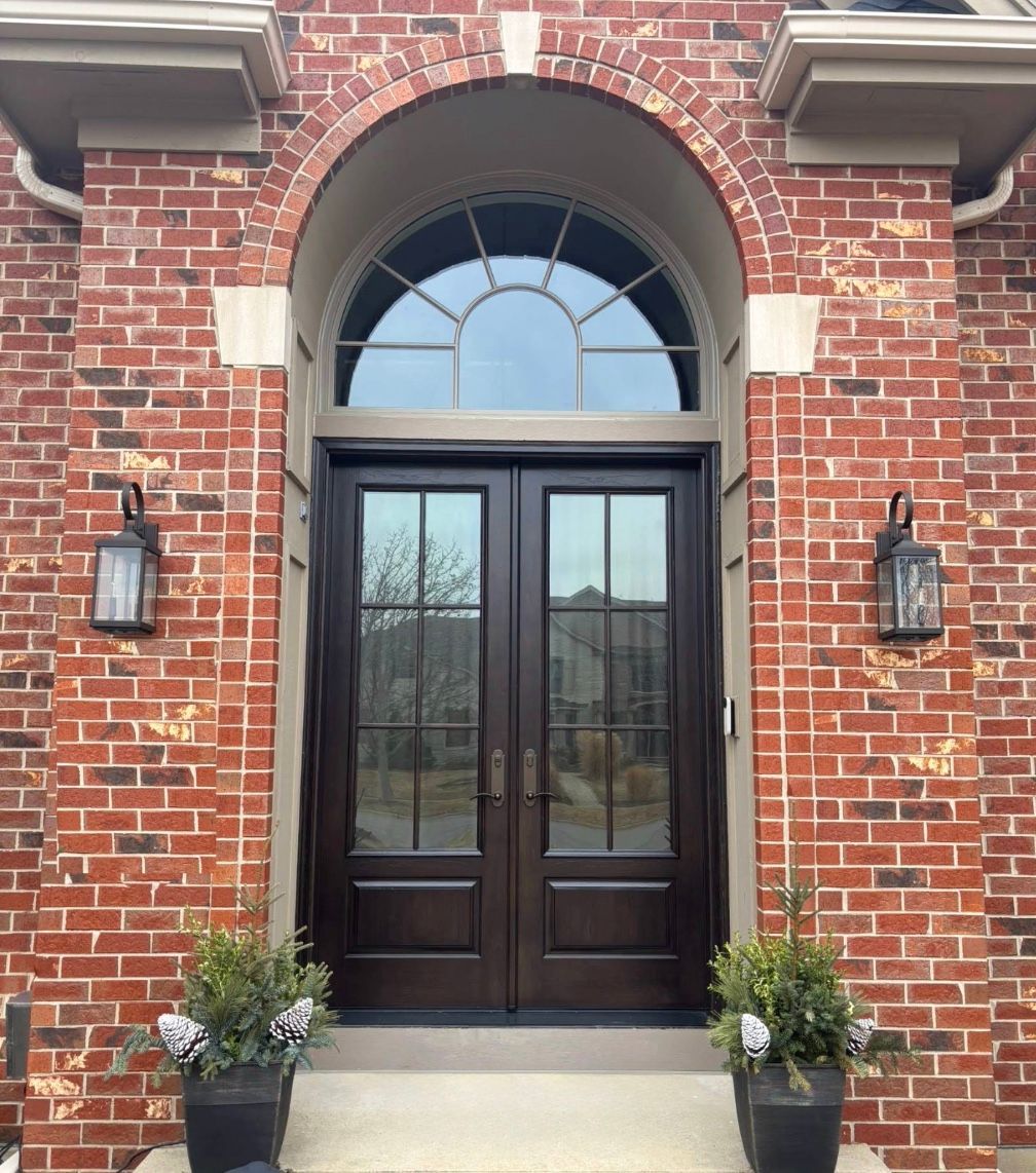 Brick facade with dark double doors, arched window, and lanterns. Potted plants flank entrance.