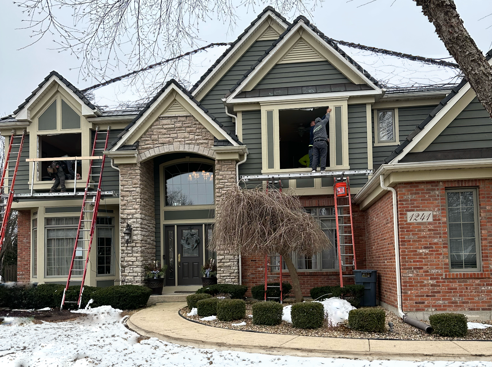 Snowy suburban house with brick and stone exterior, gabled rooflines, and red ladders set up on the front facade.