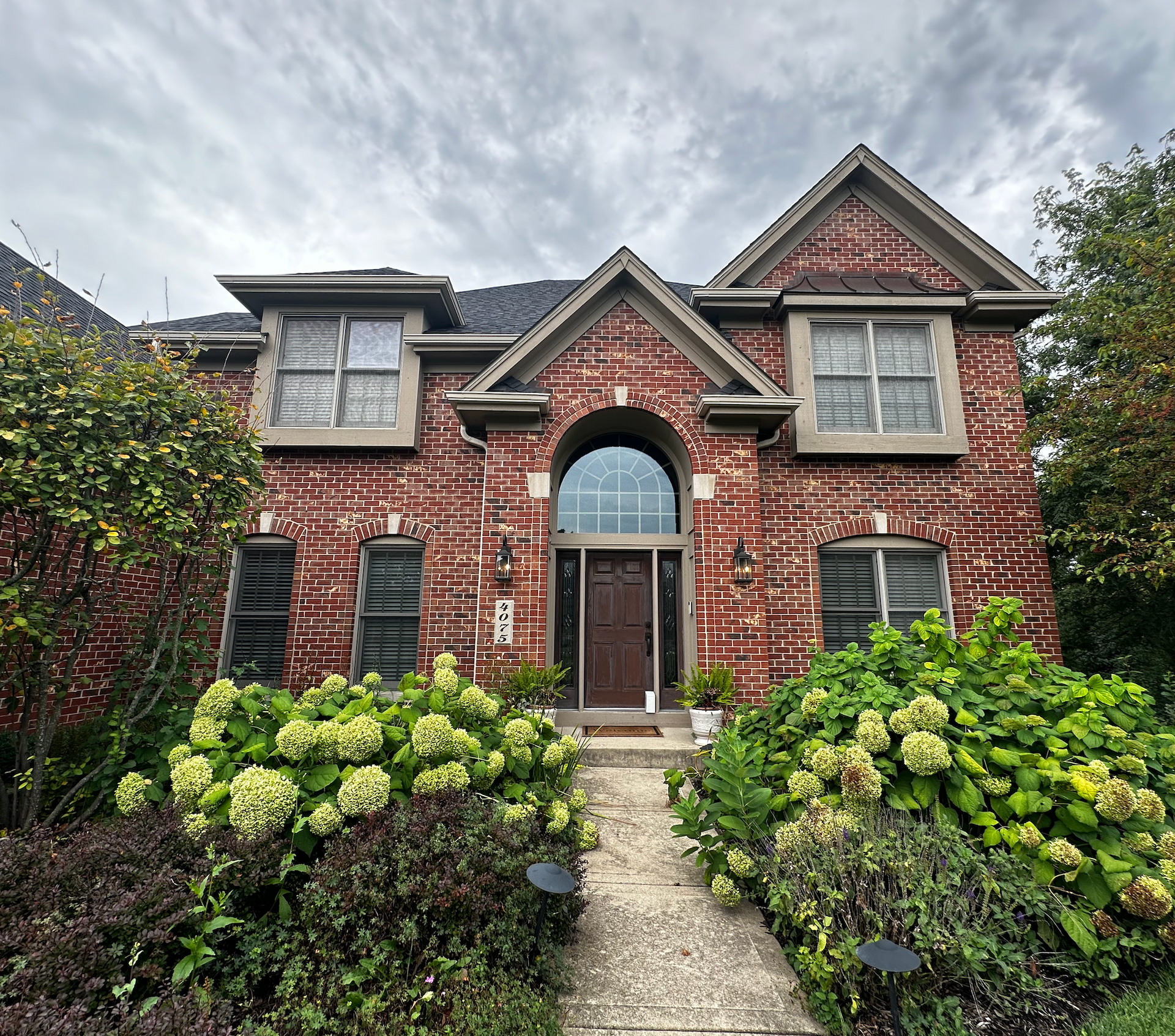 Two-story brick house with arched front door, surrounded by green bushes and a cloudy sky.