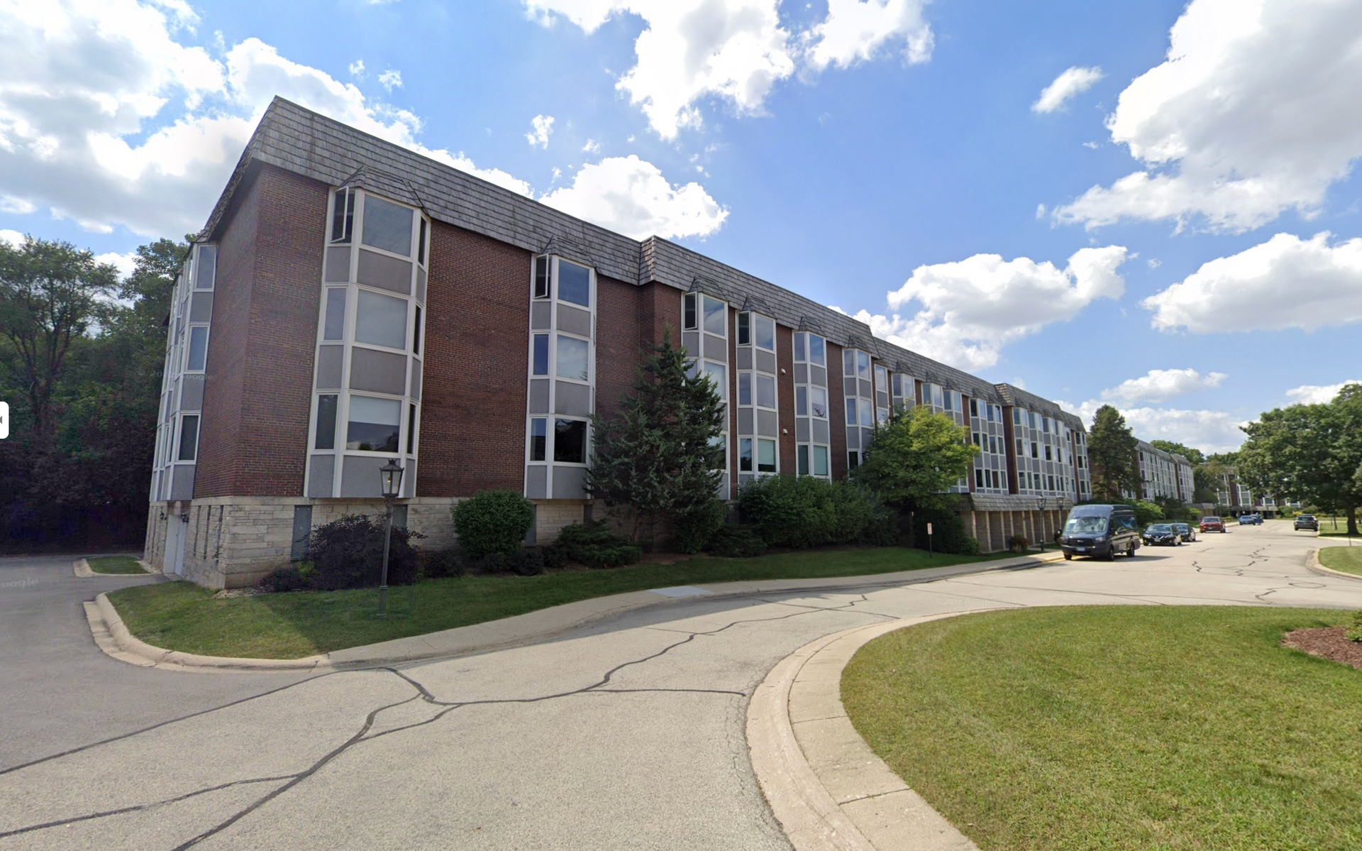Brick apartment building with angled roof and large windows; curved driveway and green lawn in front.