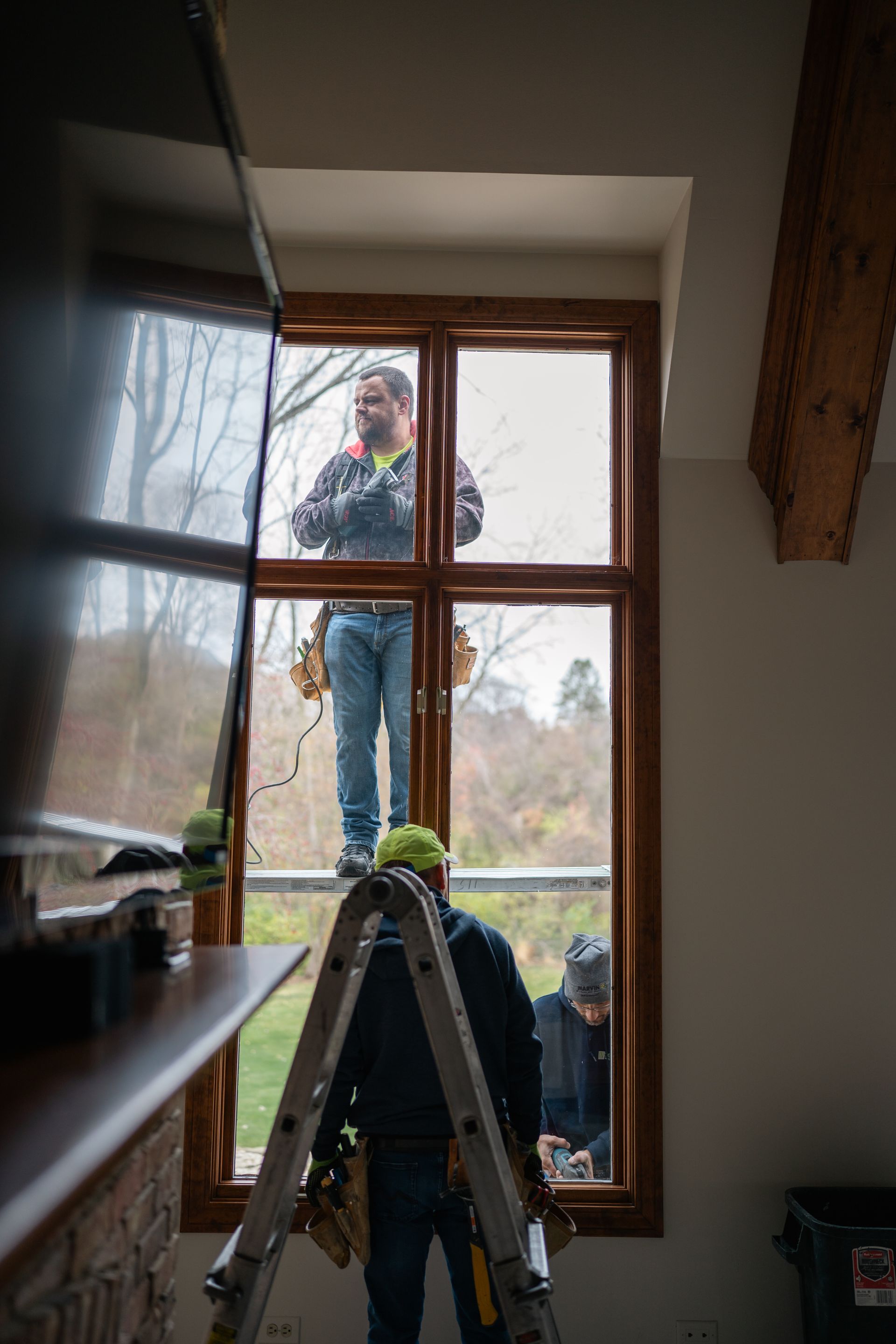 Workers installing a window inside a house; one on a ladder, two inside.