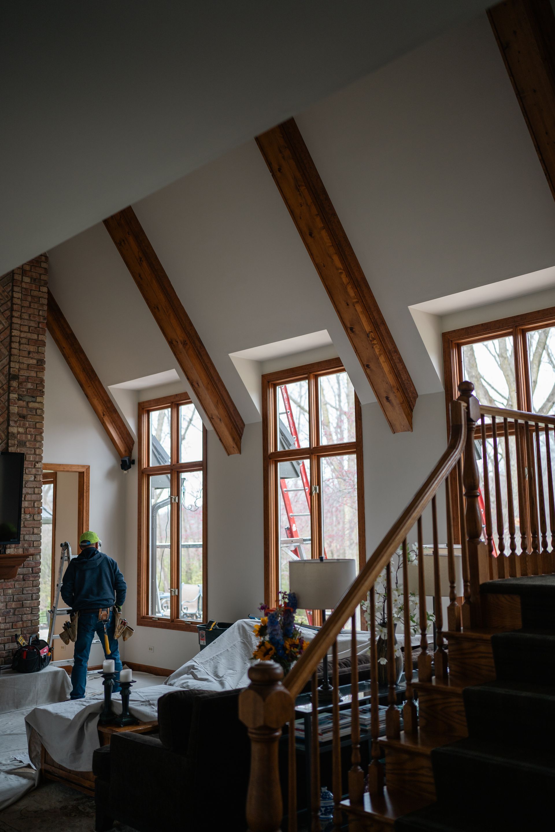 A person works in a room with wooden beams, large windows, a staircase, and a brick fireplace.