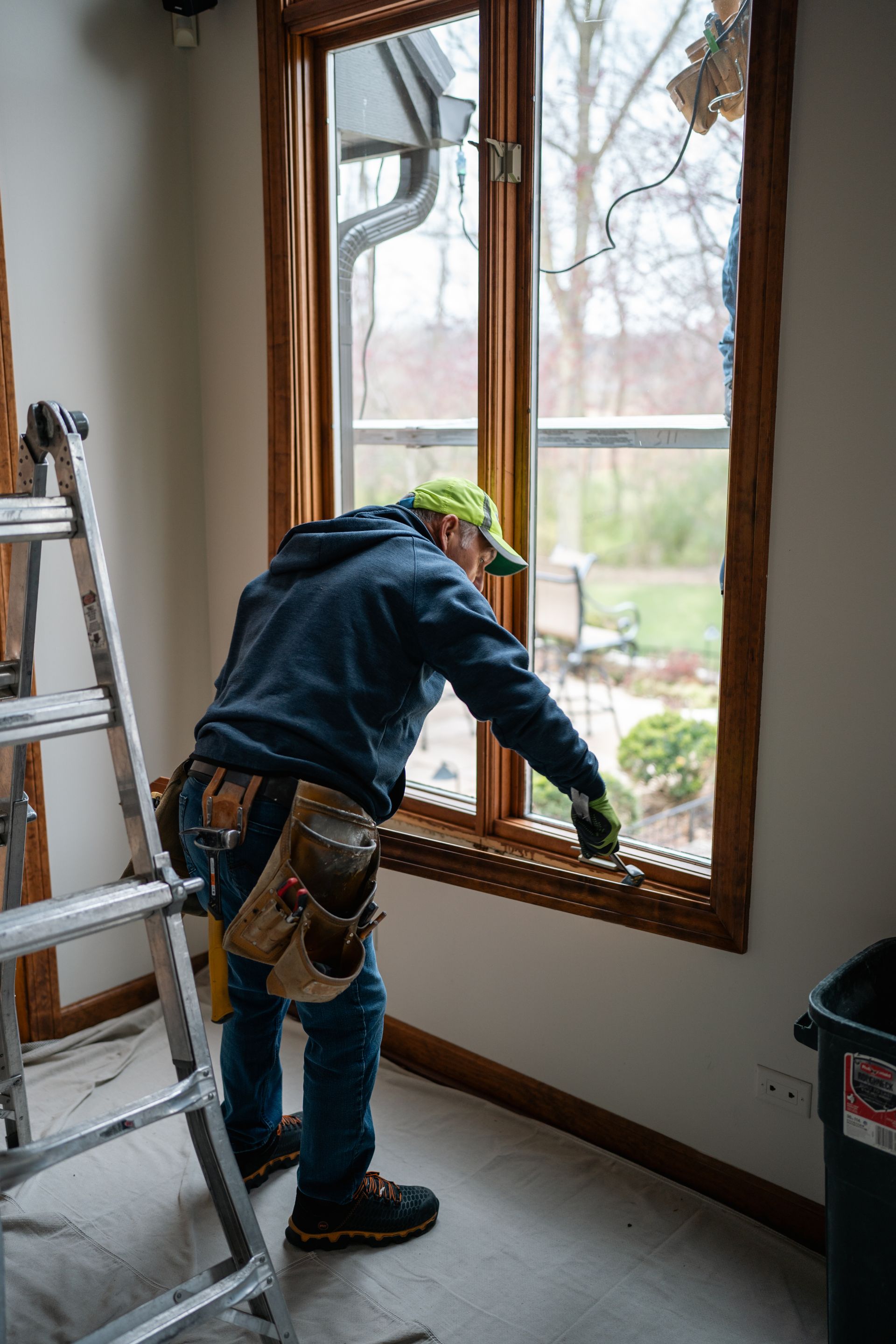 Person replacing window, wearing tool belt, next to a ladder, in a room with a view of trees.