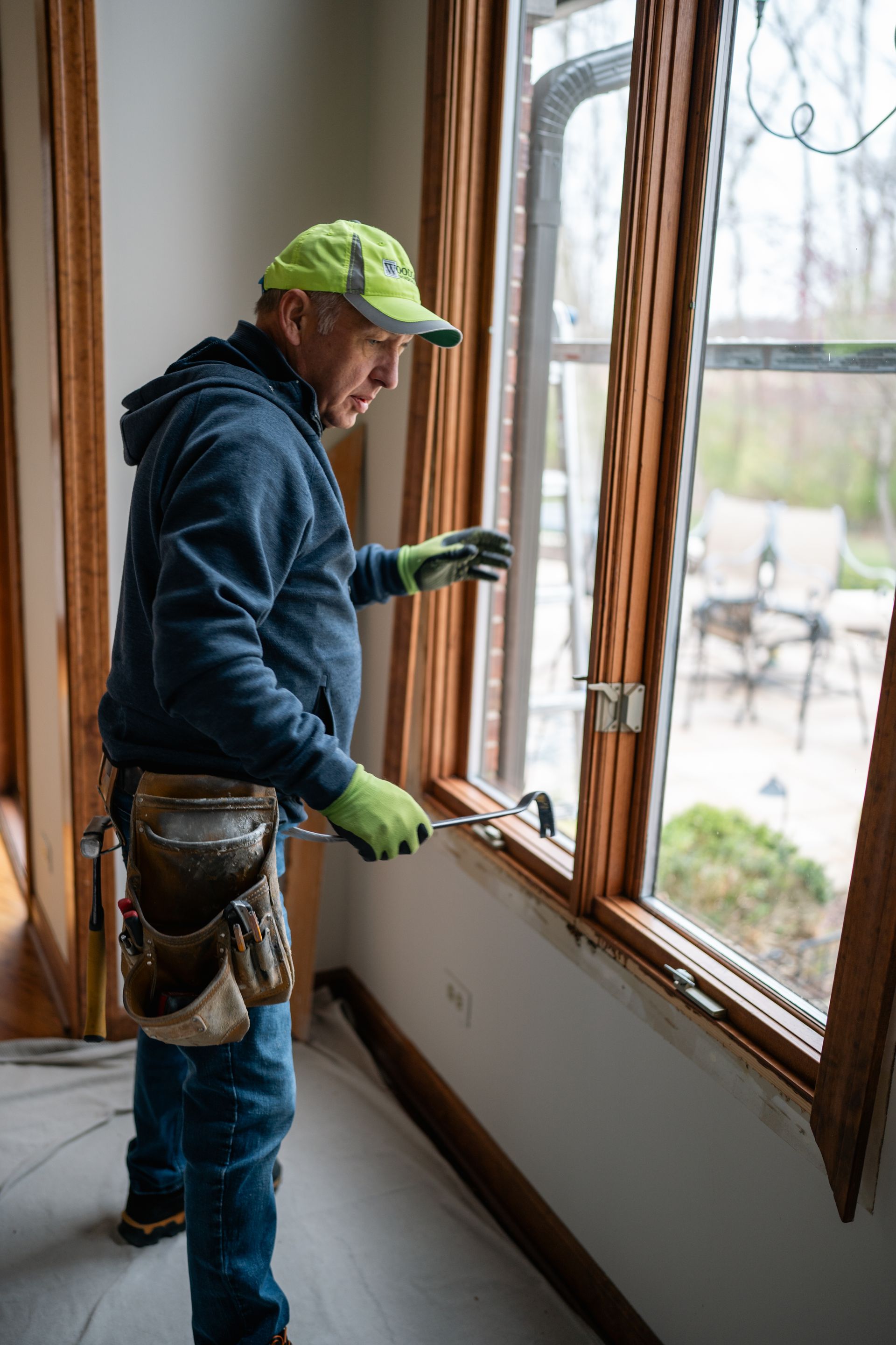 Man in work clothes replacing a window, using a tool. Indoors, next to a window, holding the frame.