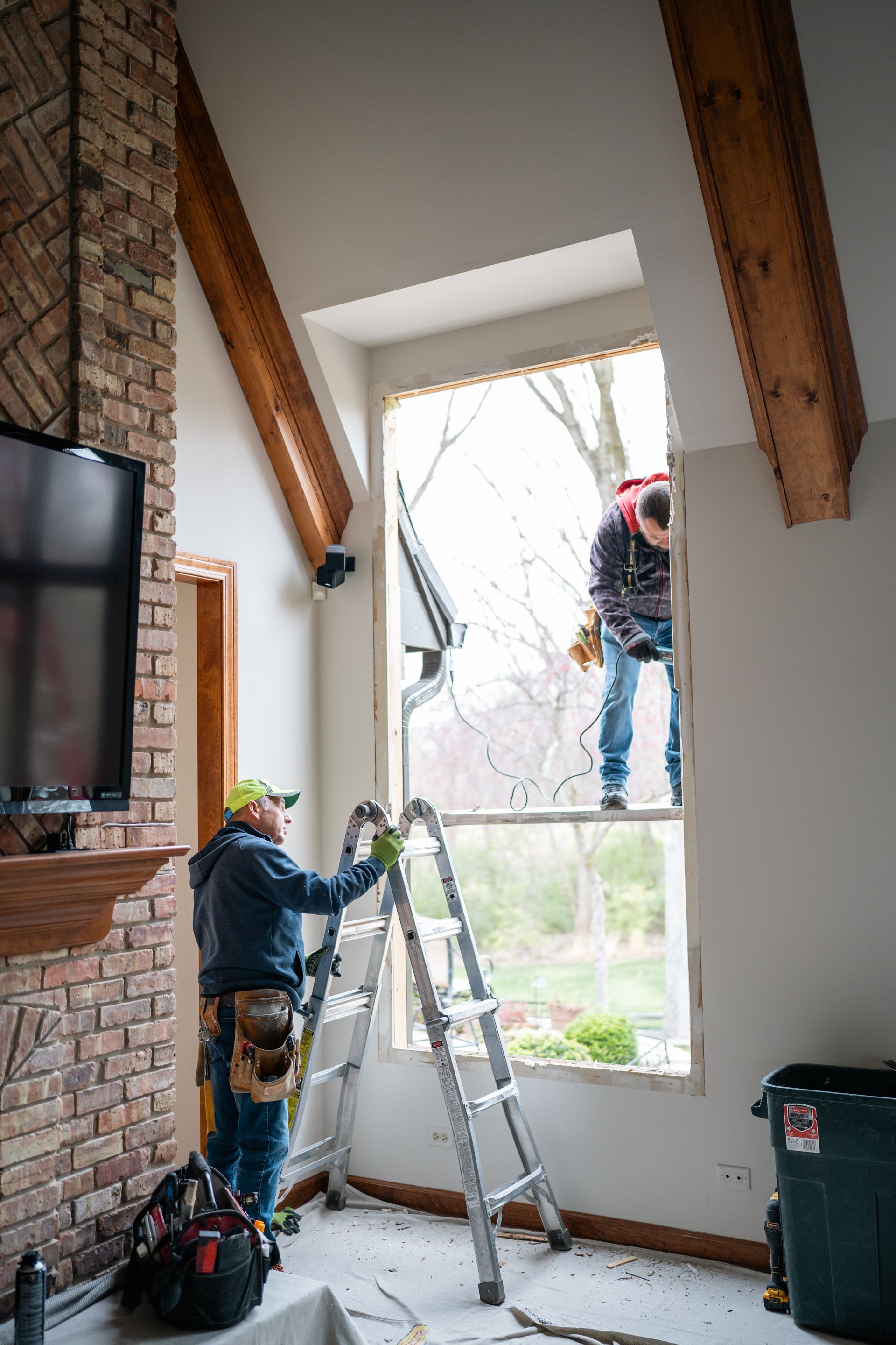 Two construction workers replacing a large window. One on ladder, one outside.