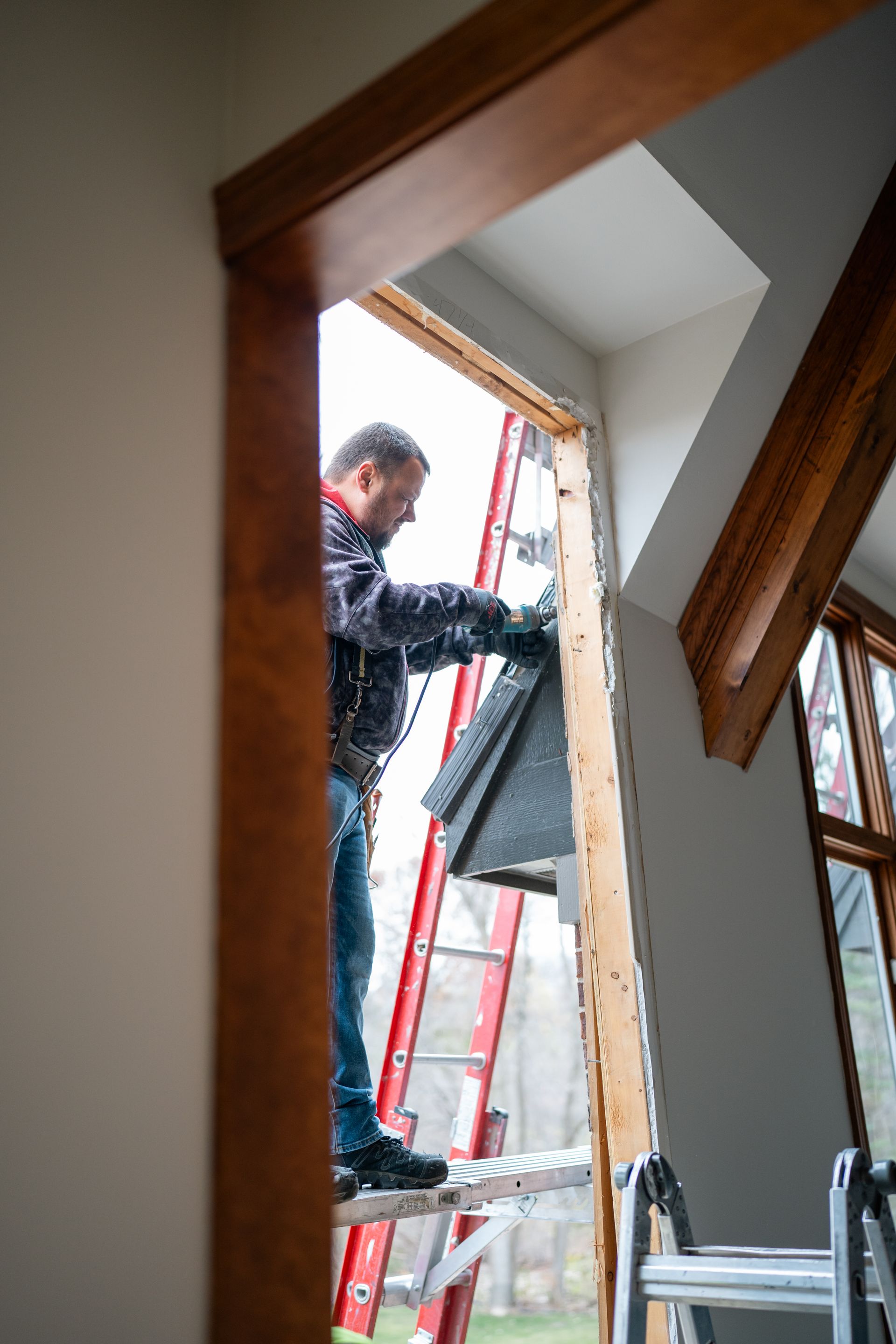 Man on a ladder installing roofing material in a window frame. Brown trim, gray shingles. Outdoor setting, overcast.