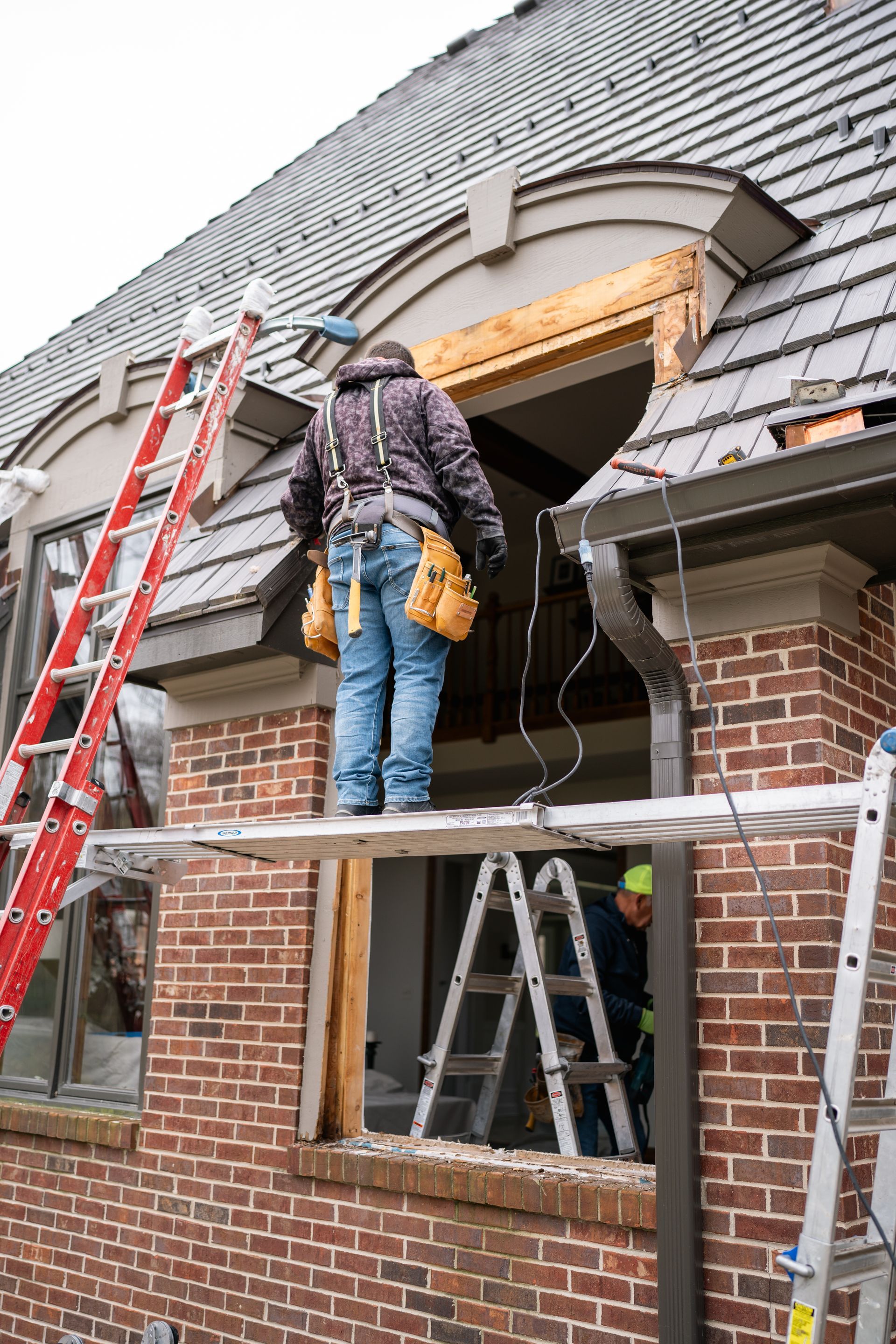 Construction workers replace a window, one standing on a scaffolding, another on a ladder. Brick building, gray roof.