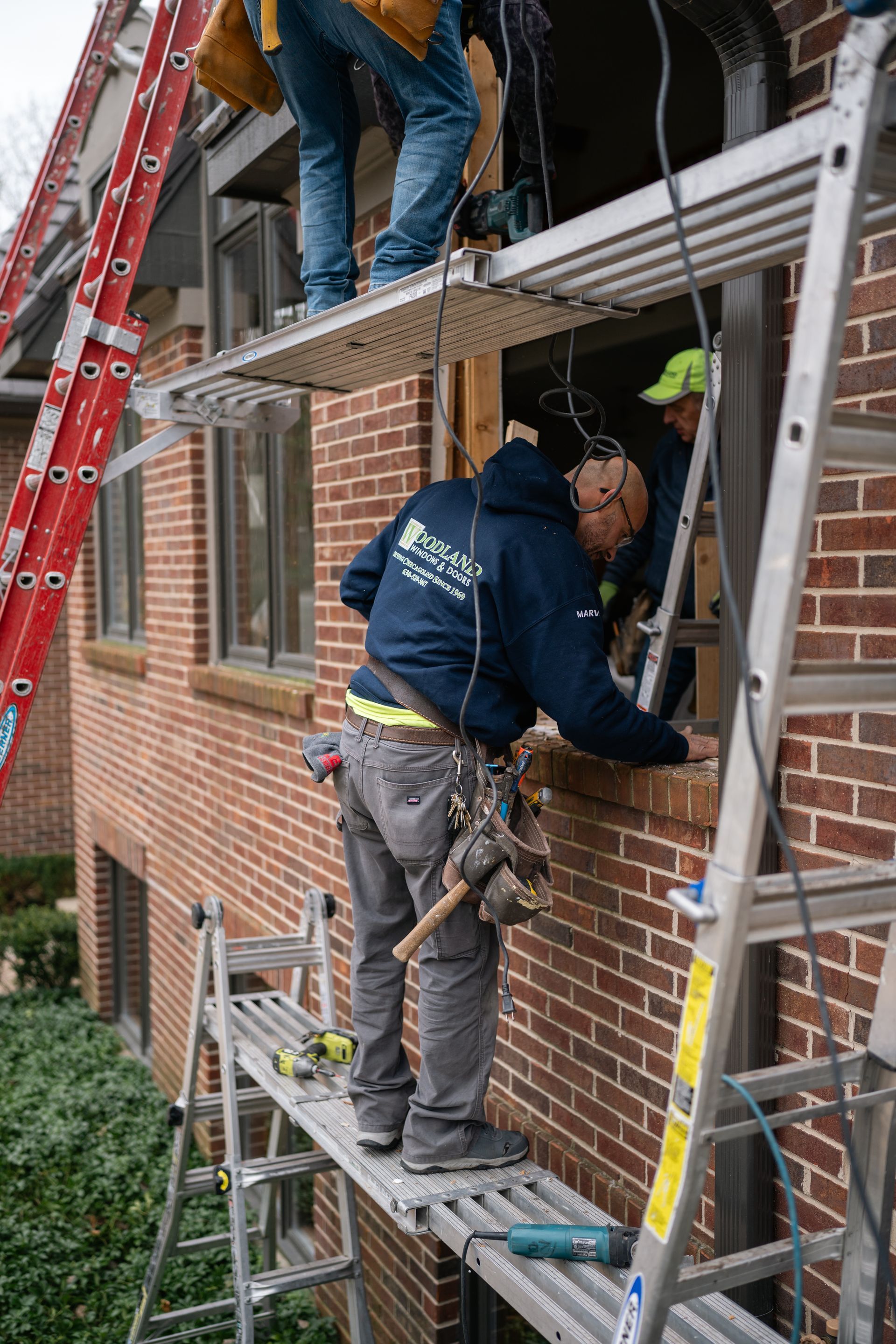 Construction workers replacing a window, using ladders and scaffolding on a brick building.