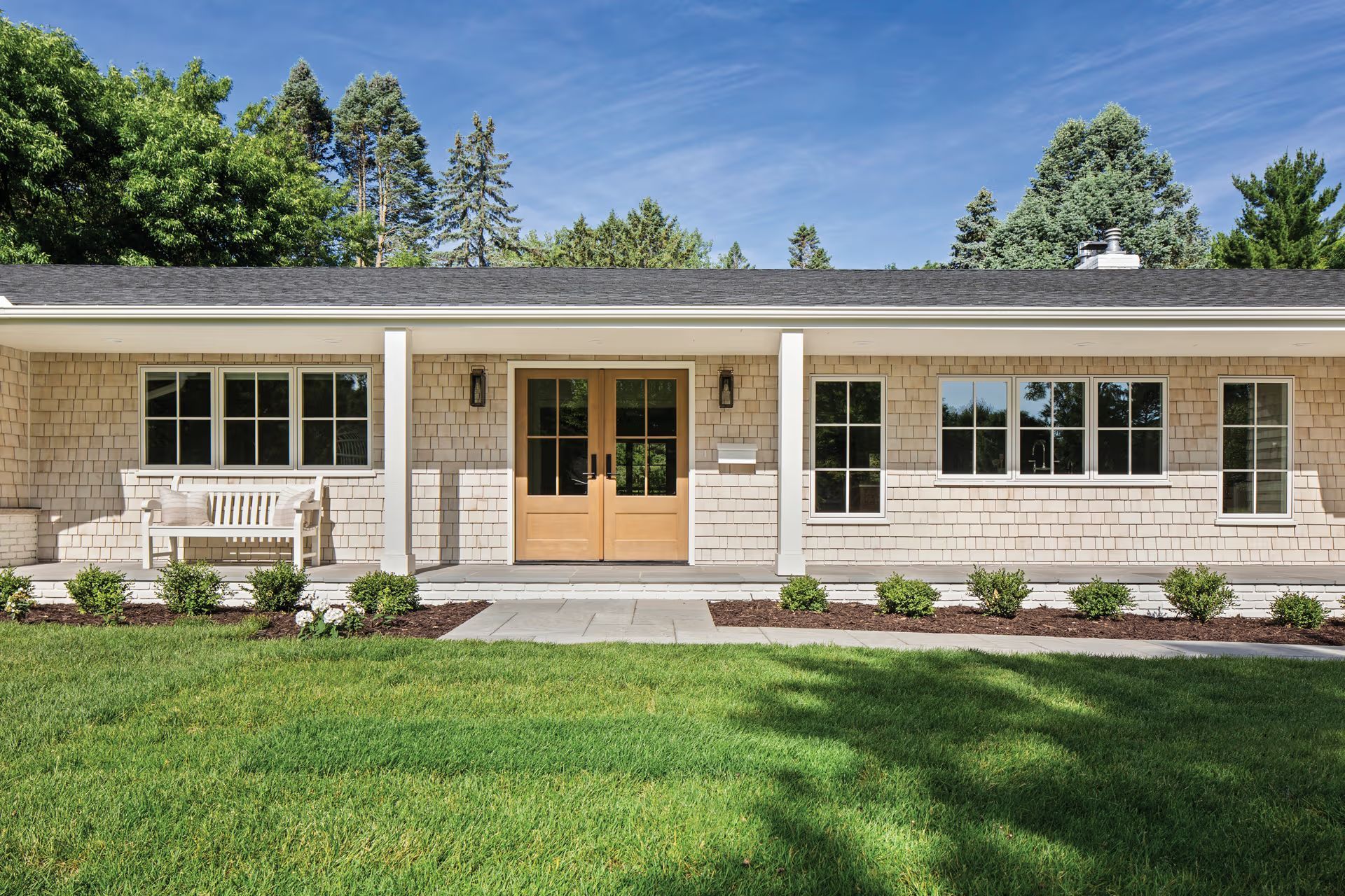 Single-story stone house with tan double doors, front porch, and green lawn under a blue sky